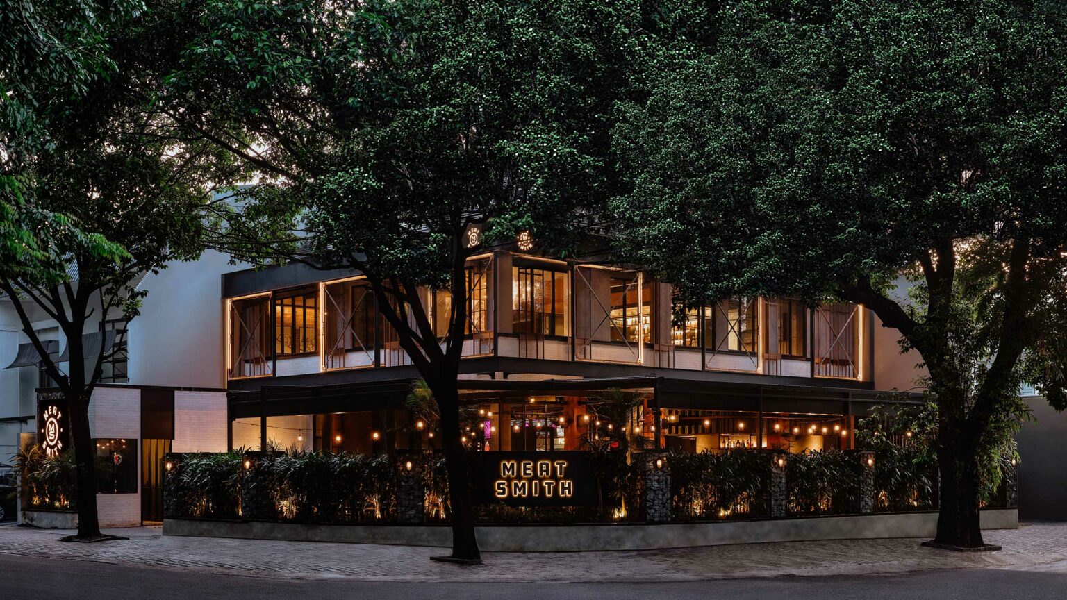 Exterior of Meat Smith restaurant, featuring a modern facade at dusk, surrounded by leafy trees. Warm lights illuminate the sign 