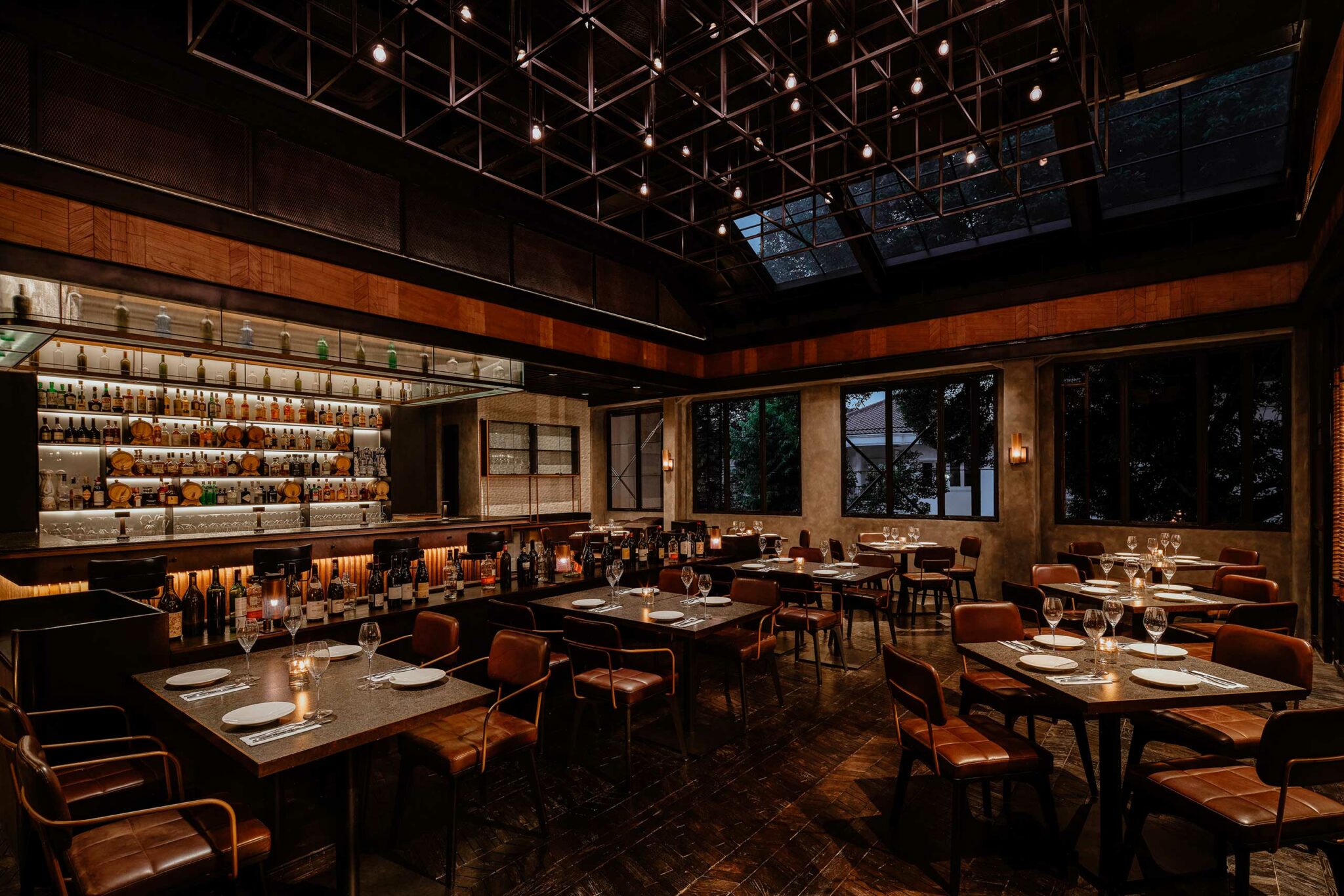 Dimly lit, elegant restaurant interior with leather chairs and square tables set with plates and glasses. A well-stocked bar and ceiling lights enhance warmth.