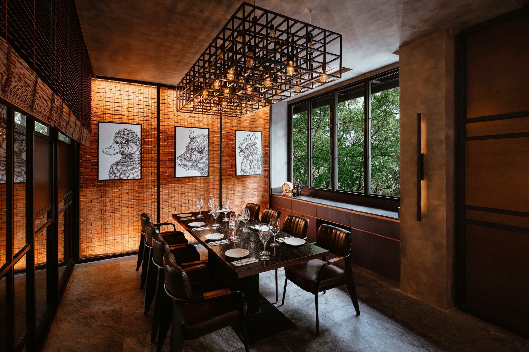 Elegant private dining room with a dark table, rich brown chairs, and stemware set. Warm lighting highlights three animal sketches on a textured wall. Large window reveals greenery, creating a cozy yet sophisticated atmosphere.