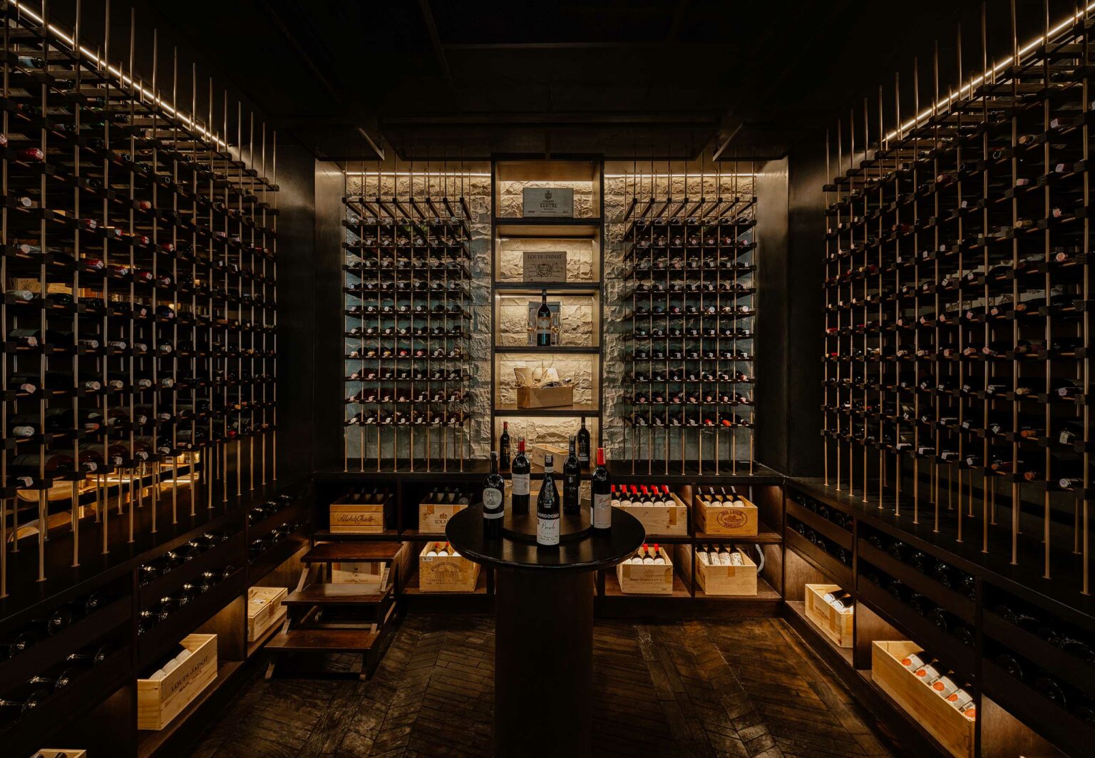 Dimly lit wine cellar with dark wood racks holding rows of wine bottles. A round table in the center displays six bottles, creating an elegant ambiance.