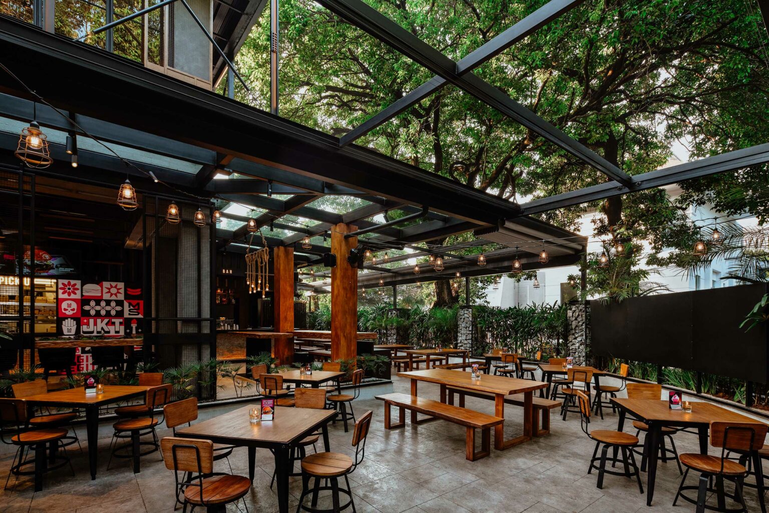 Open-air restaurant with wooden tables and chairs under a glass roof. Surrounded by lush greenery and warm lighting, creating a cozy, inviting atmosphere.