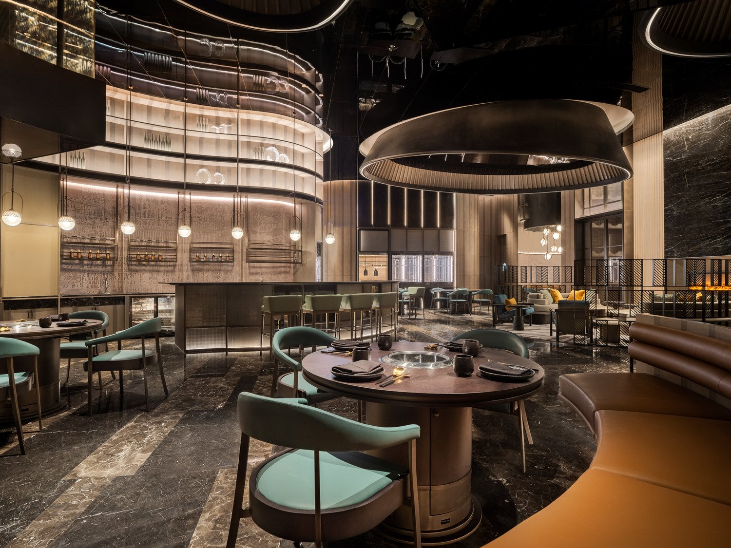 Stylish hotel lounge with a circular gray couch adorned with yellow cushions, a teal chair, facing a modern fireplace set in a black marble wall. Cozy ambiance.