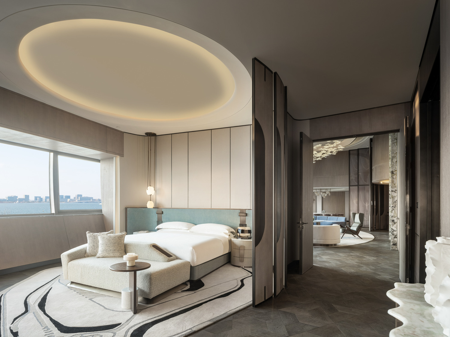 Modern suite room with elegant decor: round bed under soft lighting, marble TV wall divider, plush seating, and a small vase of flowers on a table.