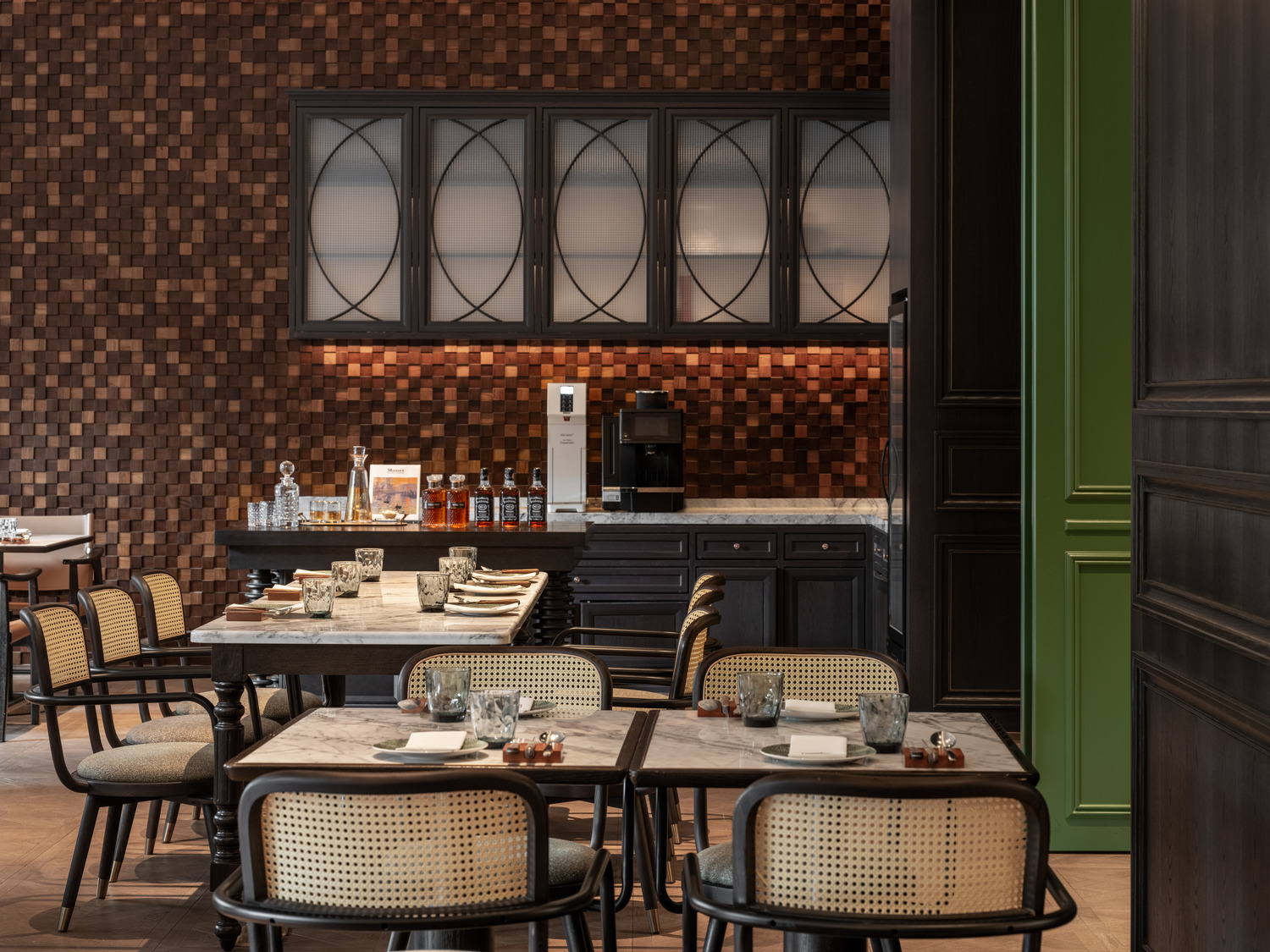 Luxurious hotel restaurant interior with geometric tile flooring, wooden chairs, and tables. Central archways lead to a floral sculpture under glass. Sophisticated ambiance.