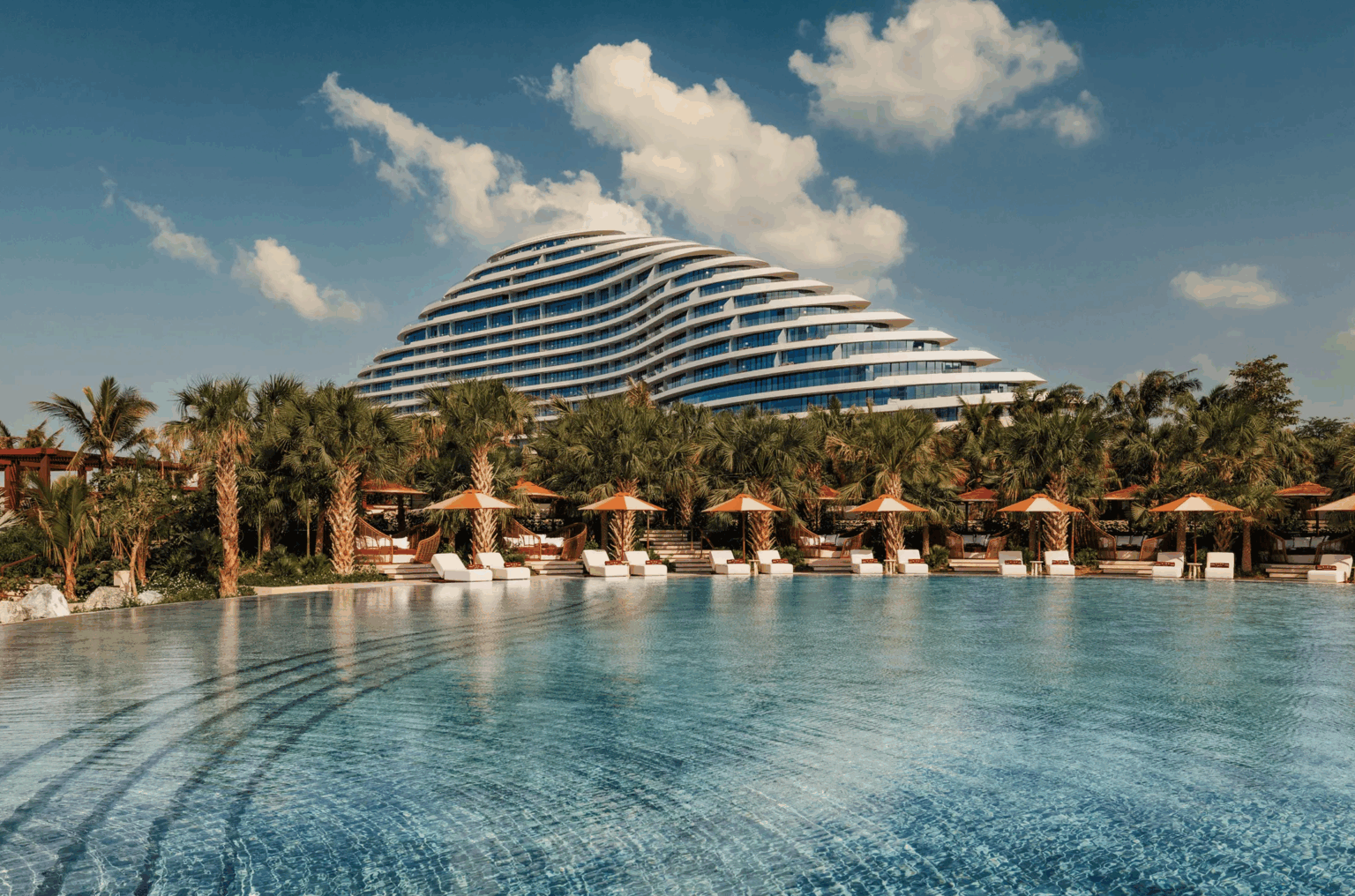 Futuristic, wave-shaped building with glass facade behind palm trees and umbrellas lining a large, tranquil pool under a clear blue sky. Luxurious and serene.