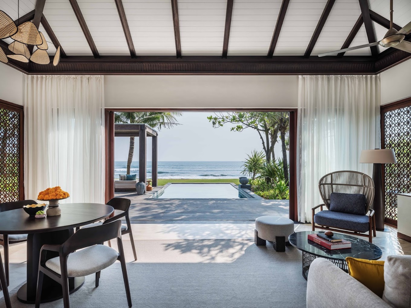 Luxurious beachfront villa interior. Open room with dark wood furniture, gentle curtains, pool leading to ocean view. Calm, inviting atmosphere.