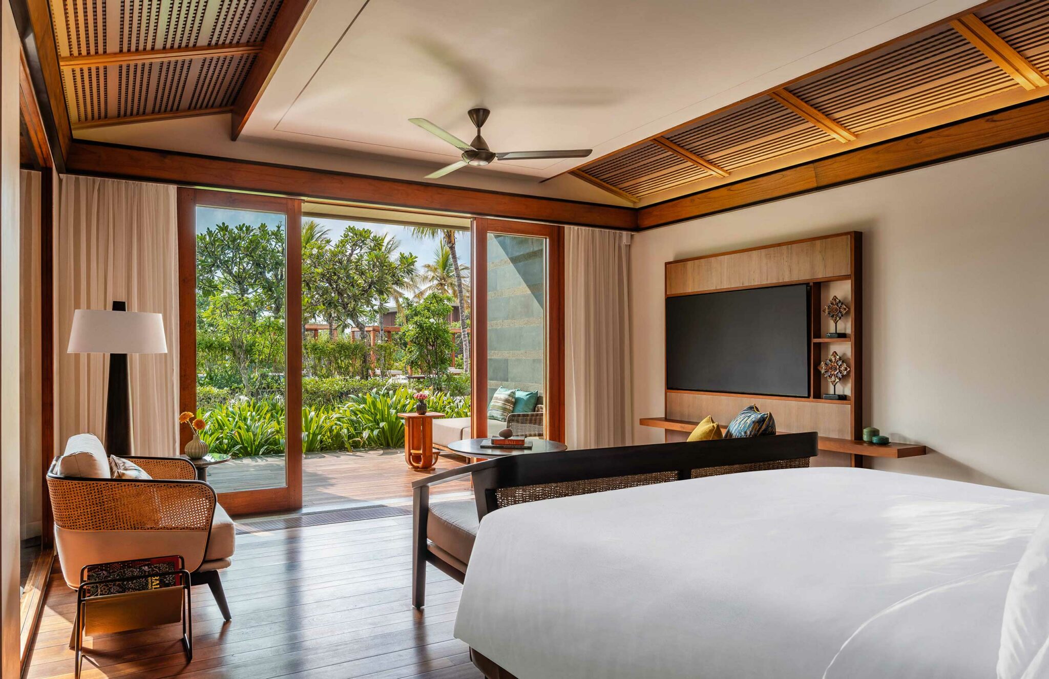 Luxurious hotel suite with a large bed, modern furnishings, and glass doors leading to a lush garden. Natural light and warm tones create a serene atmosphere.