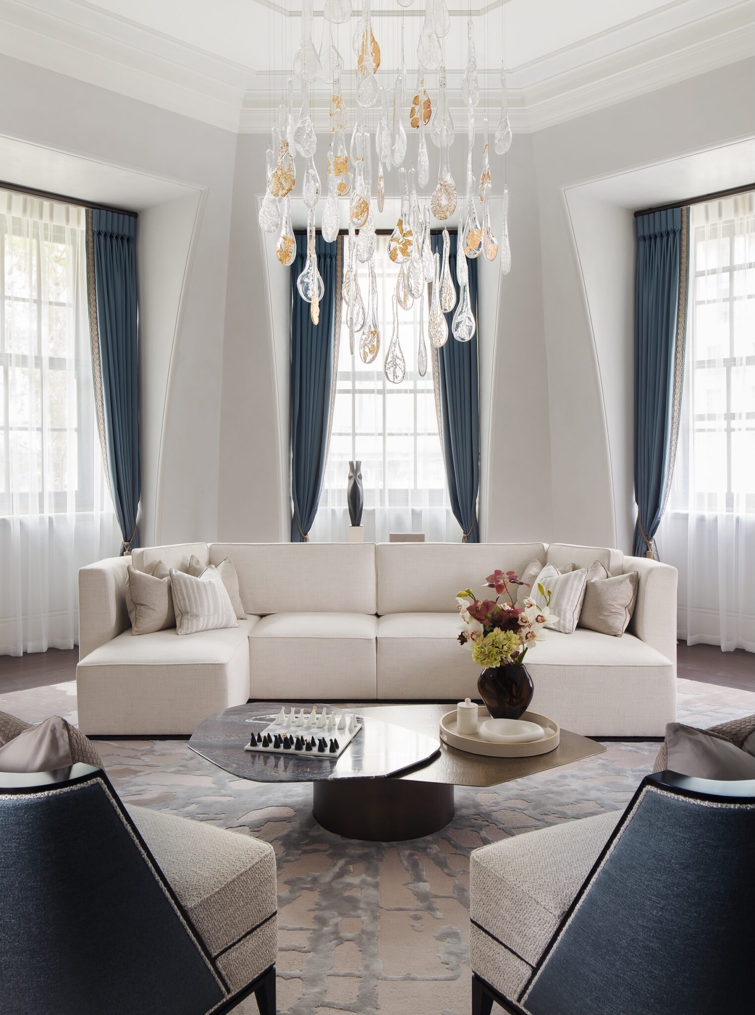 Elegant living room with a white sectional sofa, blue curtains, and a round coffee table displaying a flower arrangement. A modern chandelier adds luxury.