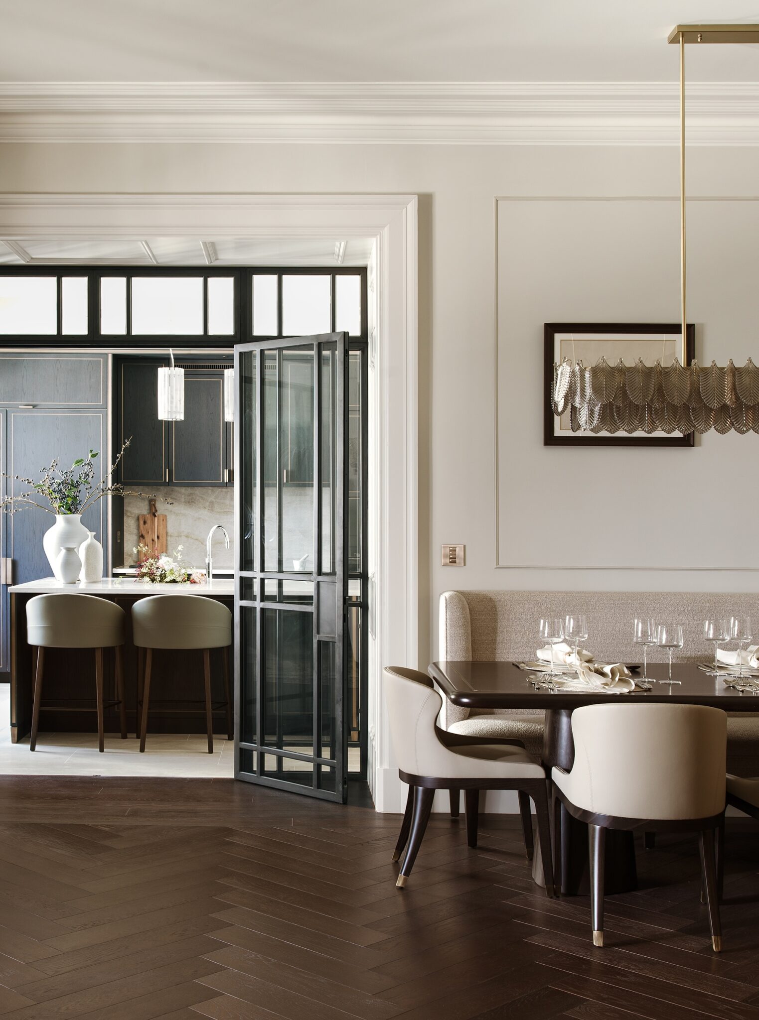 A sophisticated dining room with herringbone wood flooring, a dark wood table set for dinner, and cream chairs. An elegant modern chandelier hangs above, and black framed glass doors lead to a kitchen visible in the background.
