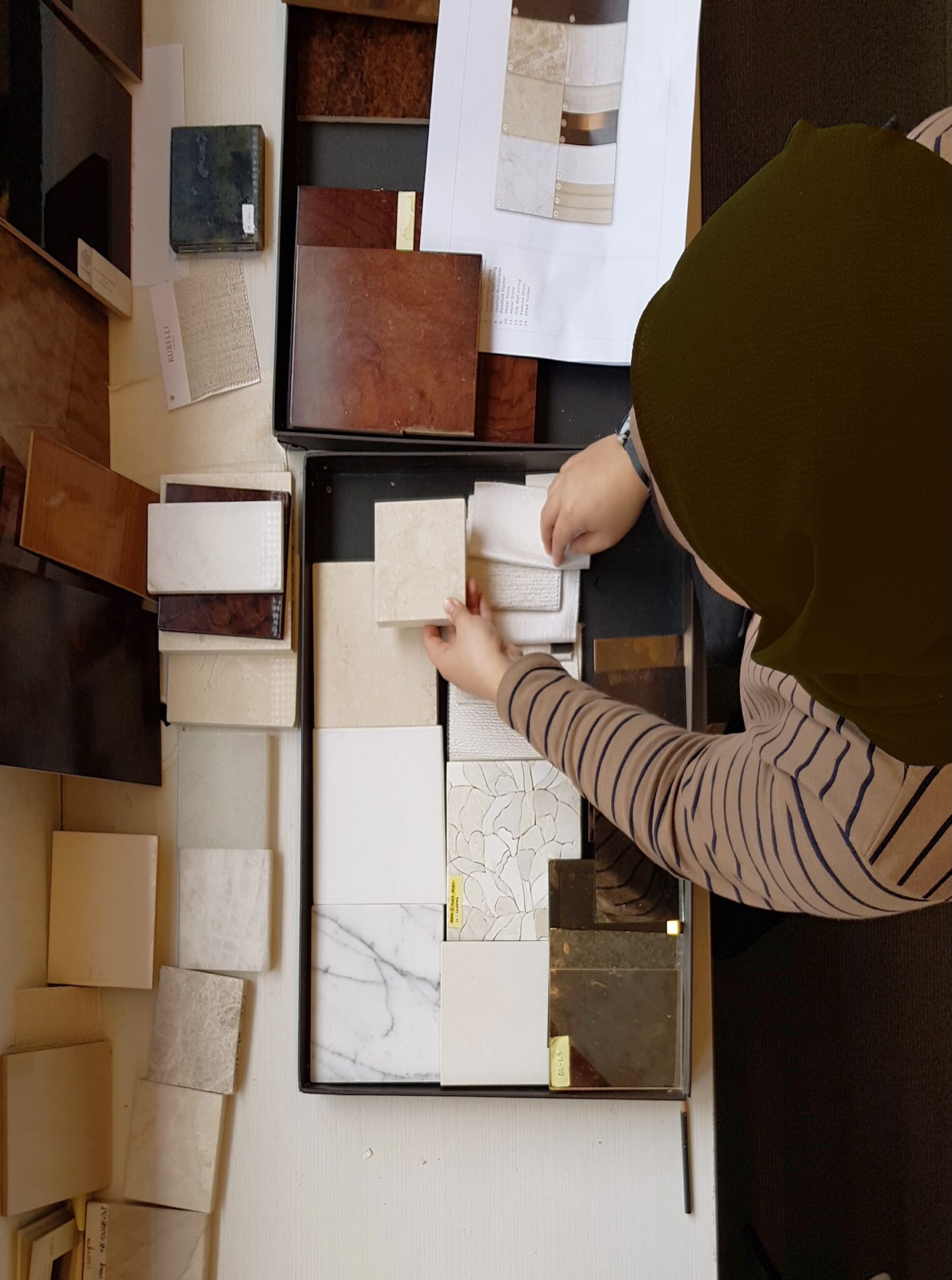 Aerial view of a young female designer in striped shirt organizing an assortment of tile samples in different colors and textures on a table, conveying thoughtfulness and creativity.