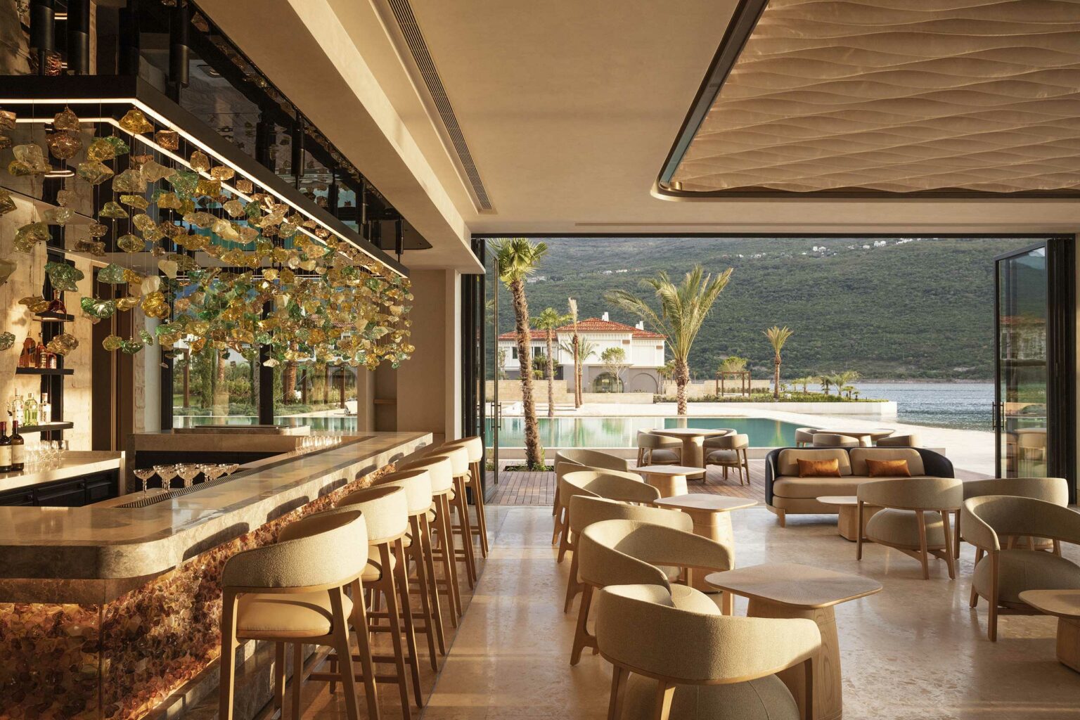Luxurious open-air bar with elegant bar and modern seating. Sunlight streams through, revealing a pool with palm trees and scenic mountain view.