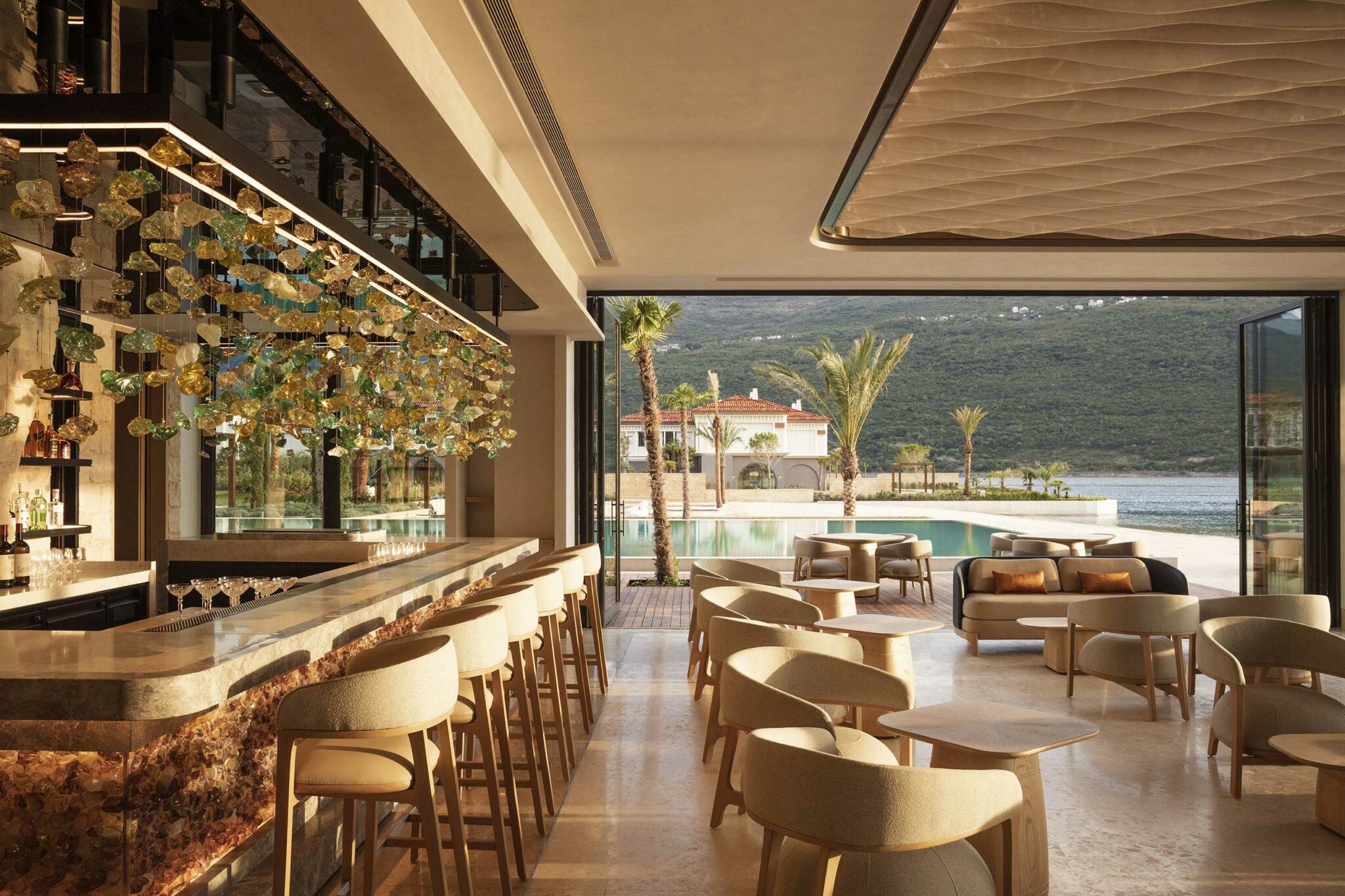 Luxurious open-air bar with elegant bar and modern seating. Sunlight streams through, revealing a pool with palm trees and scenic mountain view.