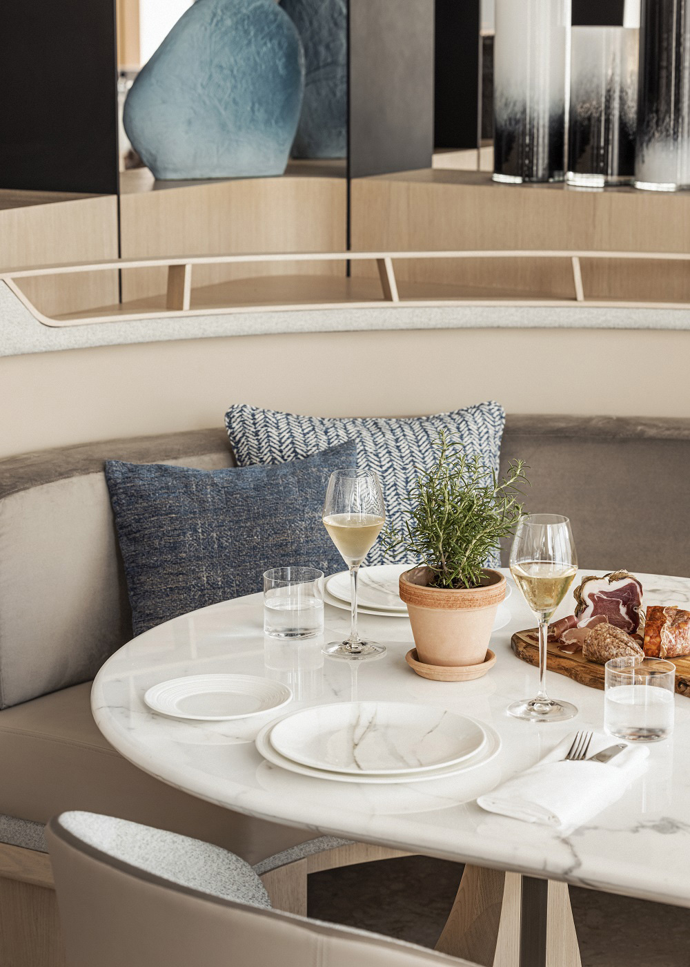 Cozy dining setting with a marble table, two glasses of white wine, potted herb, and charcuterie. Soft blue pillows and modern decor create a relaxed atmosphere.