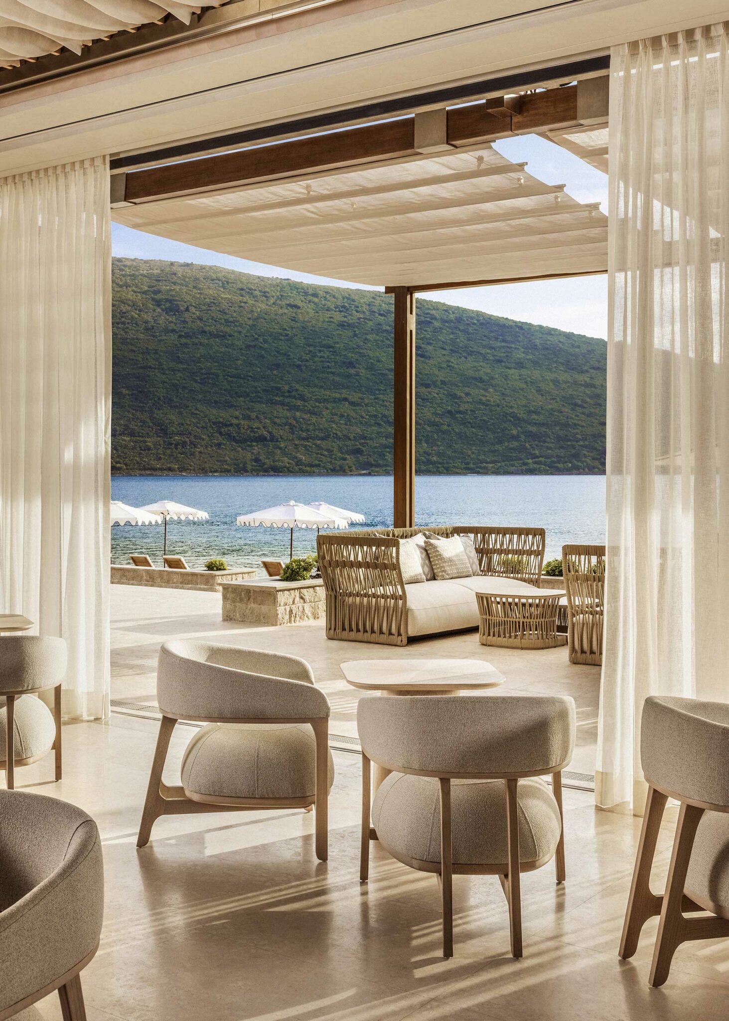 Luxurious open-air lounge with elegant chairs and sofas facing a serene blue sea and lush hillside. White curtains and umbrellas create a tranquil ambiance.