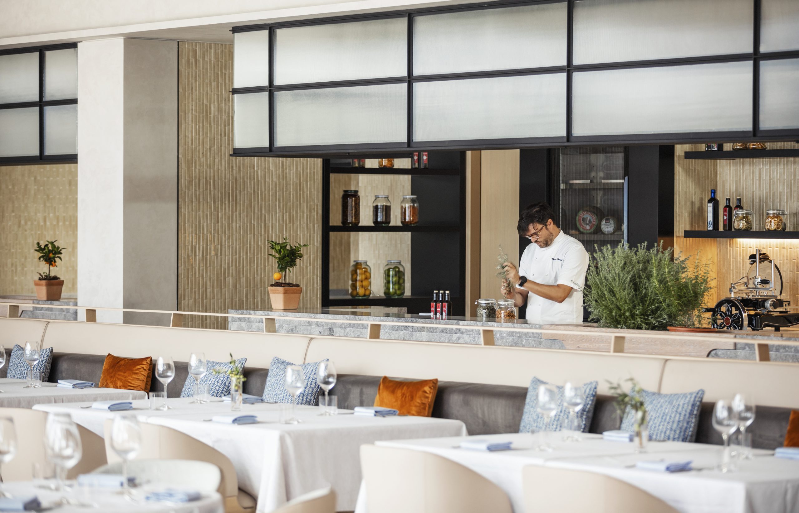 A chef in a modern, elegant restaurant kitchen garnishes a dish. Tables with white cloths, wine glasses, and vibrant cushions fill the foreground.