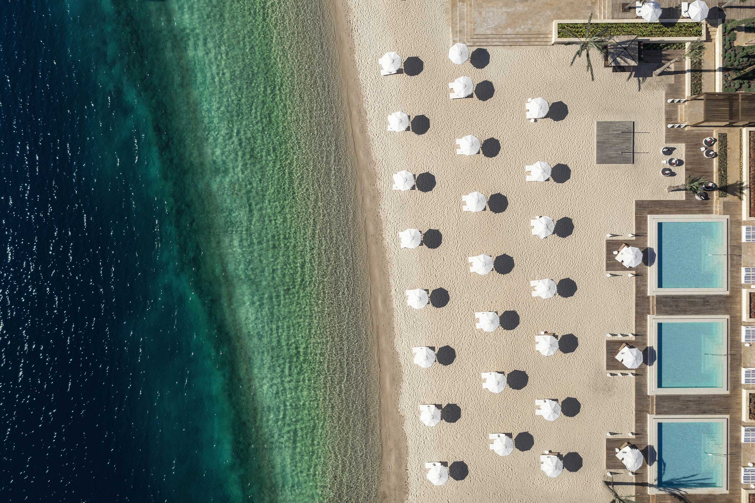 Aerial view of a serene beach with rows of white umbrellas and loungers on golden sand, adjacent to three turquoise pools, creating a tranquil resort ambiance.