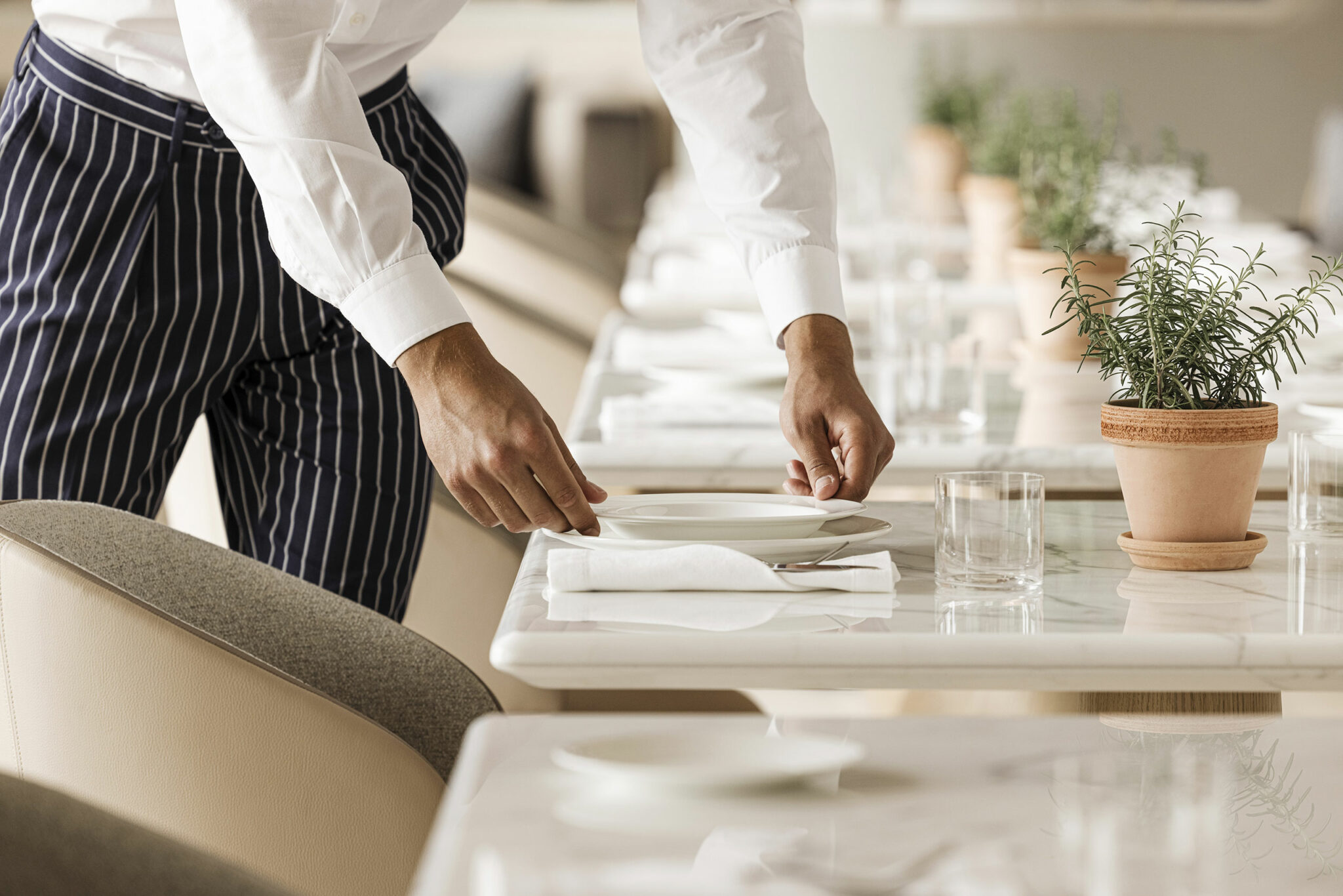 A restaurant server in a white shirt and striped pants meticulously sets a table with plates, folded napkins, and glasses, while potted plants add a fresh touch.