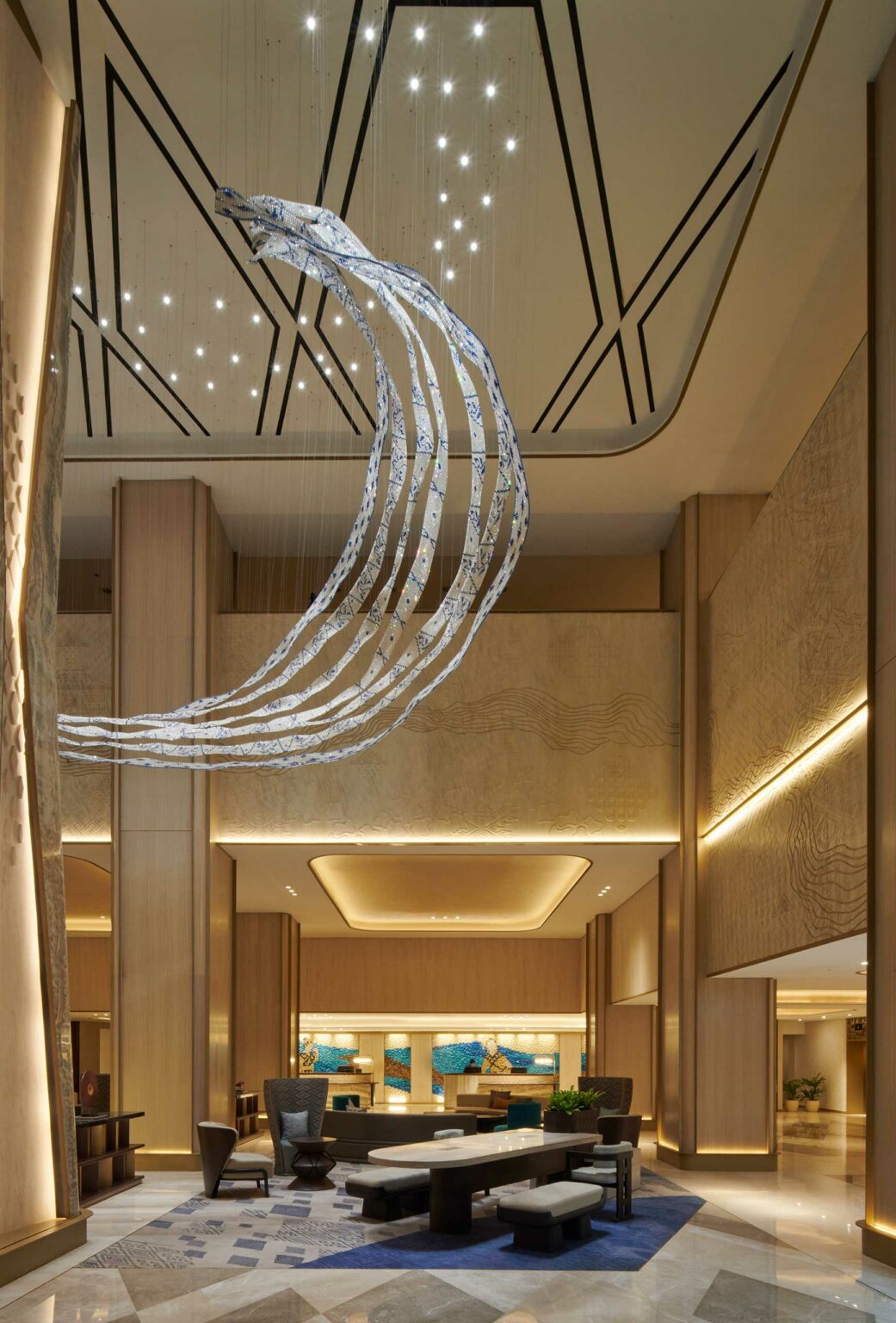 Spacious hotel lobby with a large, curved metal sculpture hanging from the ceiling. The room features warm lighting and contemporary wooden decor.