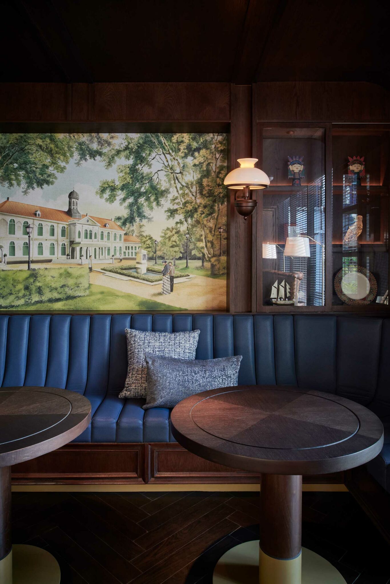 Cozy cafe interior of a hotel with blue cushioned seating, two circular wooden tables, and decorative pillows. A large painting of a grand building and trees adds elegance. Warm lighting enhances the inviting atmosphere.