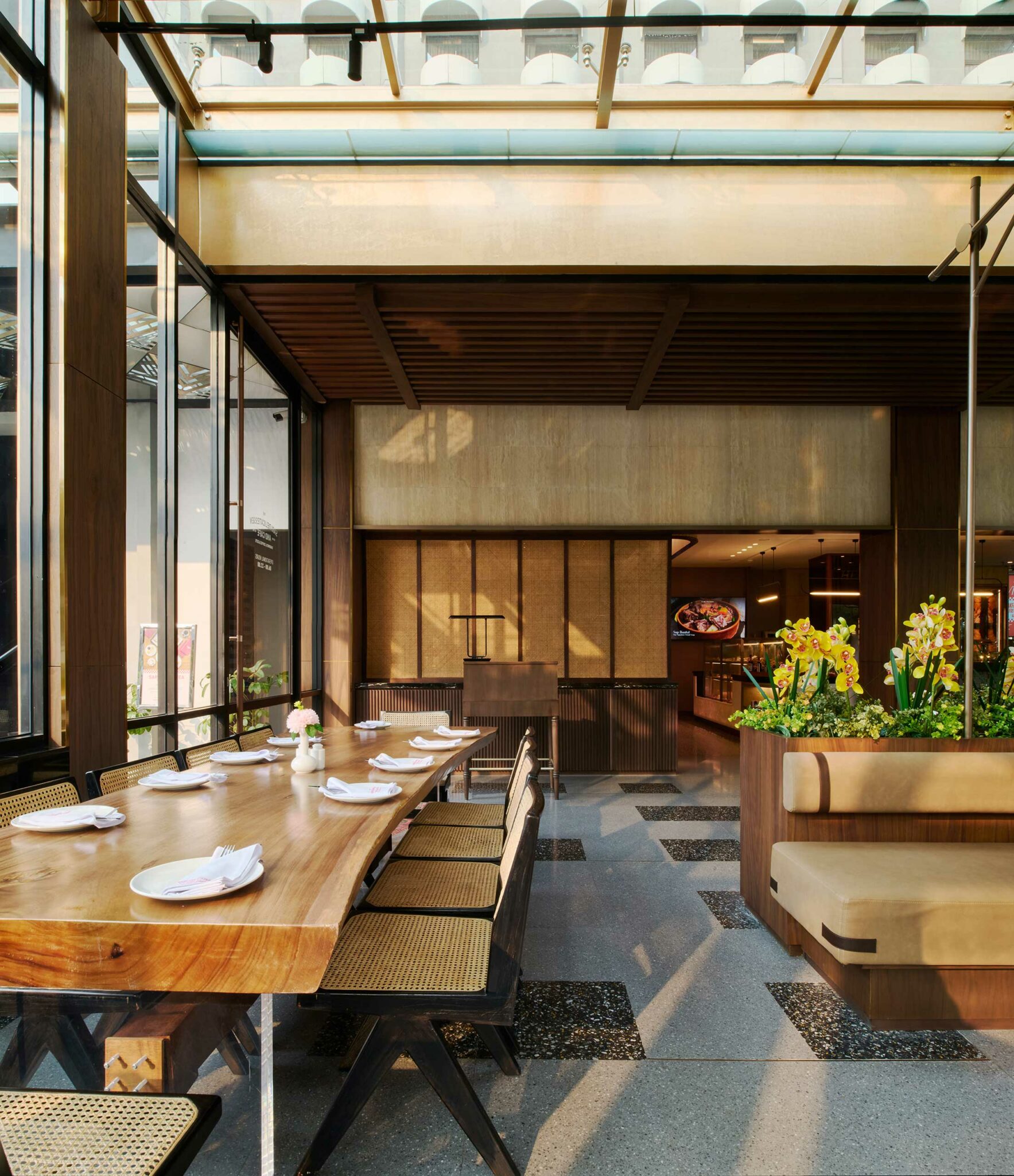 Spacious hotel all day dining area featuring a long wooden dining table set with neatly folded napkins, surrounded by wicker chairs, with sunlight streaming in through large windows and decorative flowers adding a lively touch.