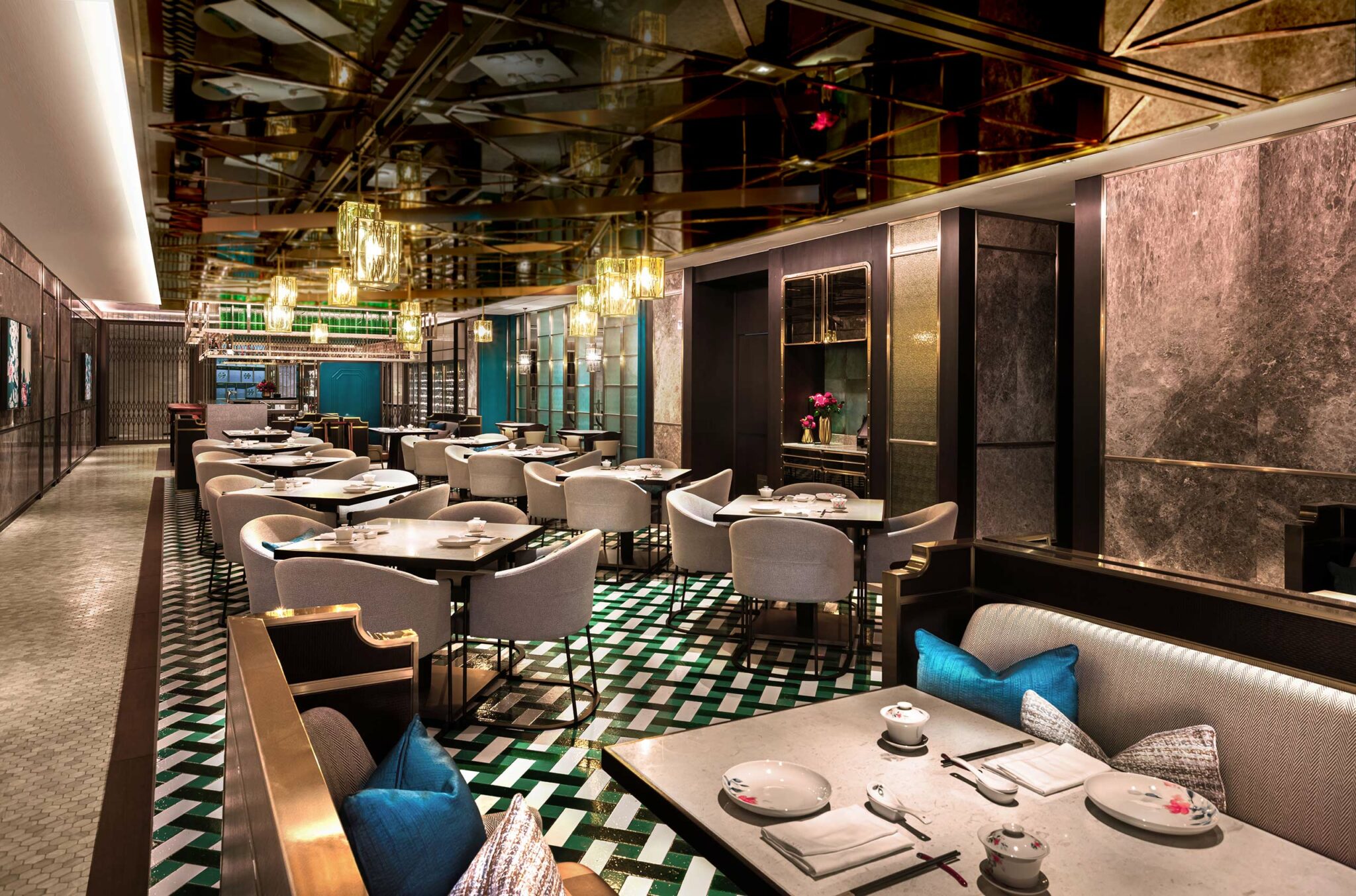 Elegant restaurant interior with modern decor, featuring patterned green and white flooring, stylish grey chairs, and warm lighting. Tables set for dining.