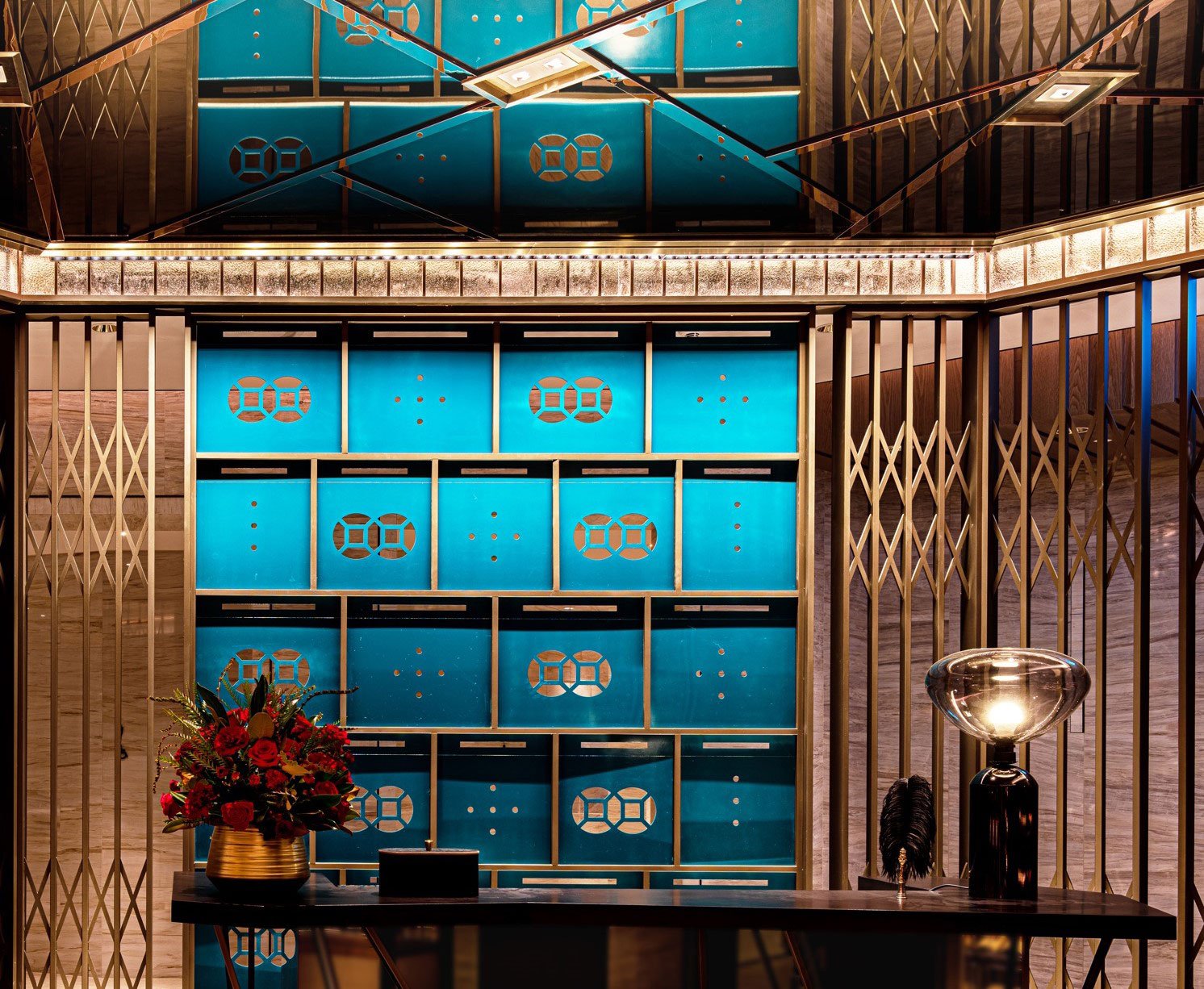 Elegant hotel restaurant reception with teal accents, geometric wall patterns, warm lighting, and red floral arrangement creating a welcoming ambiance.