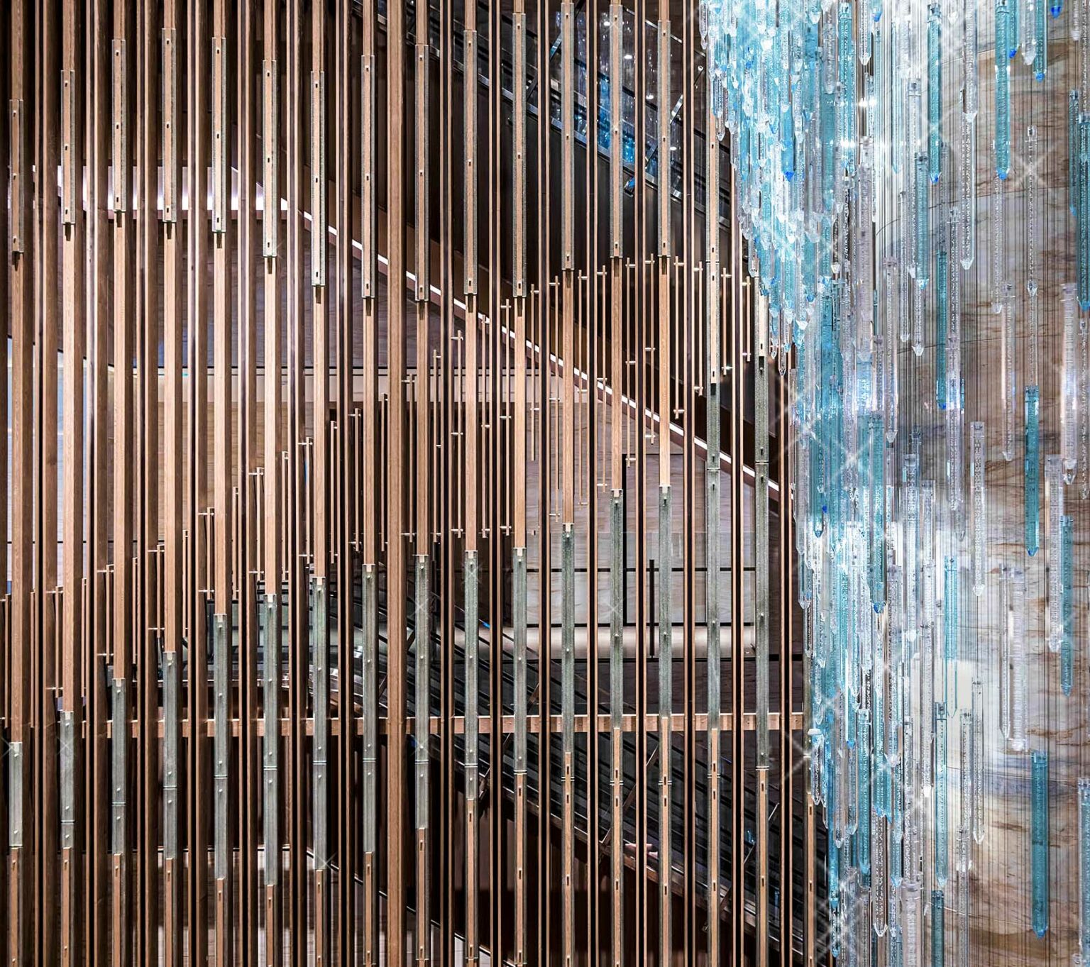 Hotel lobby chandelier close-up with vertical wooden slats creating geometric patterns alongside a cascading installation of transparent blue crystals, conveying elegance.