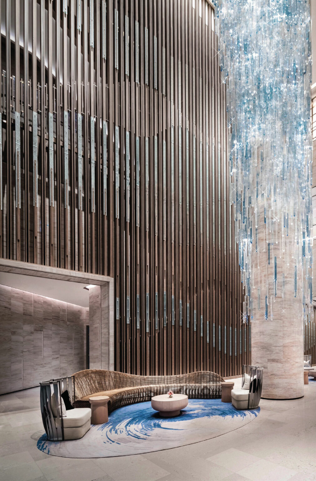 Luxurious hotel lobby with a grand, cascading glass chandelier. Curved seating on a blue and beige carpet creates an elegant, modern atmosphere.