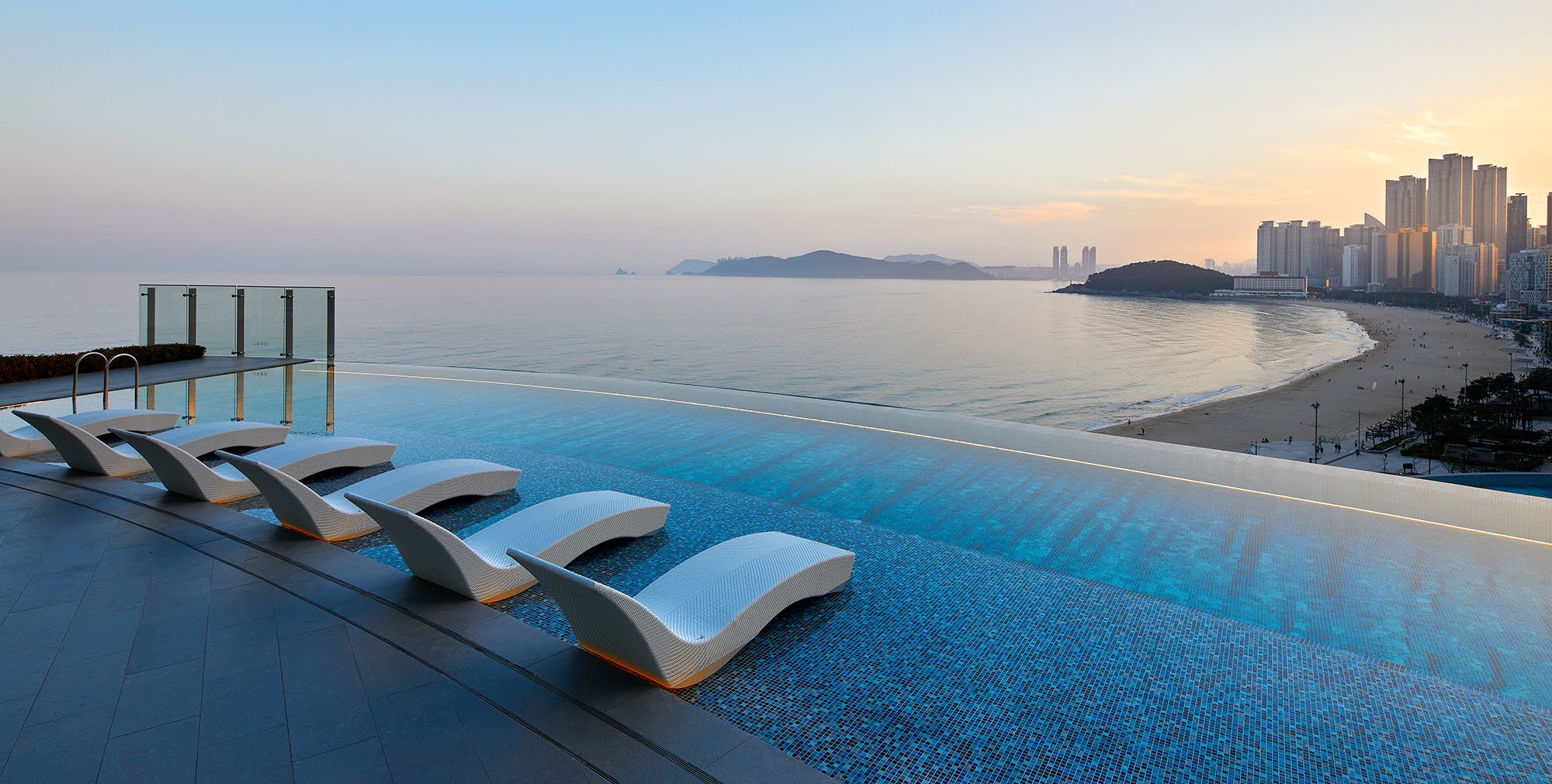Infinity pool with sleek white loungers overlooks a serene beach, calm ocean, and distant Busan's skyline at sunrise, creating a tranquil atmosphere.