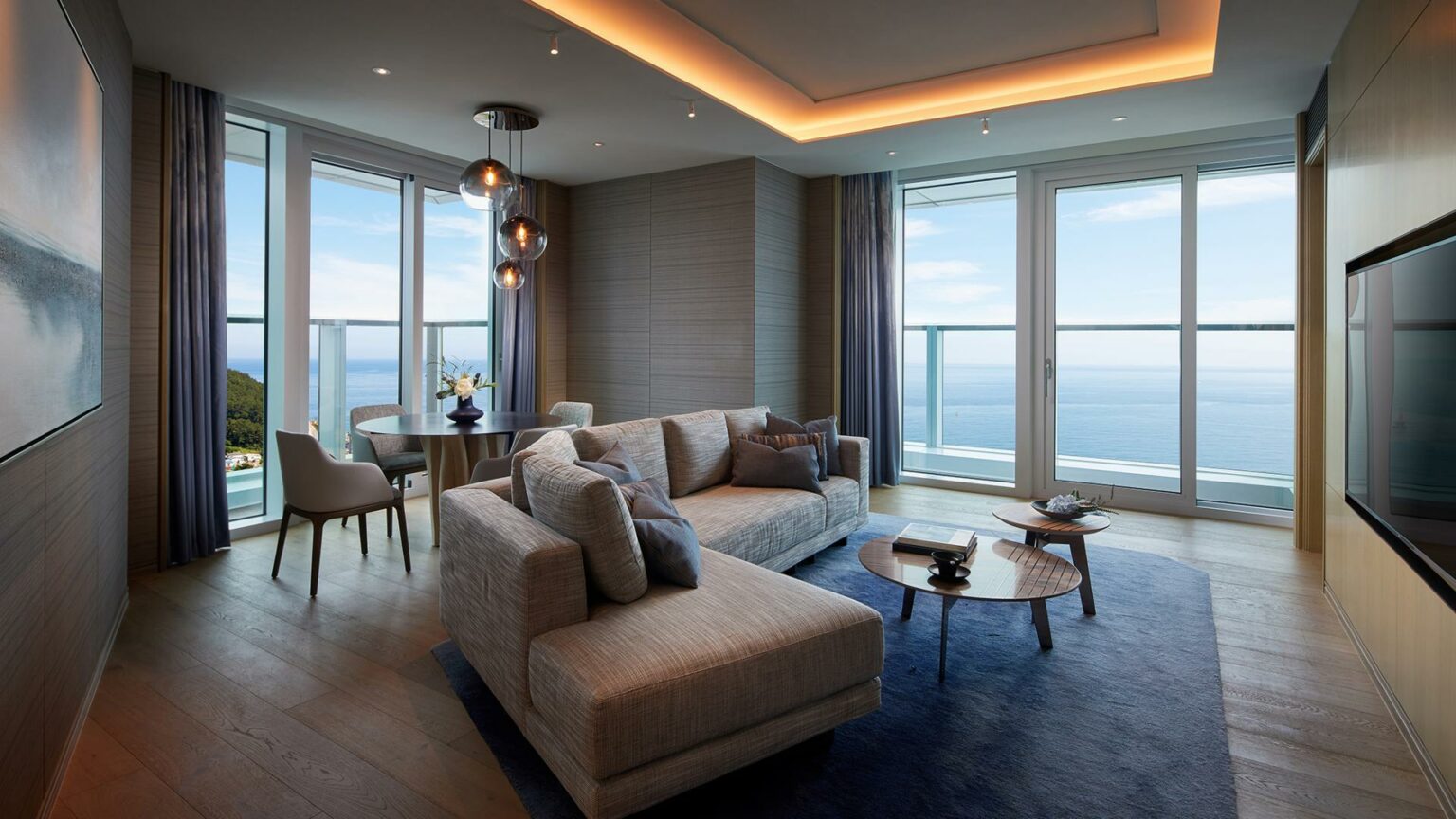Modern guest room living room with ocean view, featuring a beige sectional sofa, blue rug, and round coffee table. Floor-to-ceiling windows create a serene atmosphere.