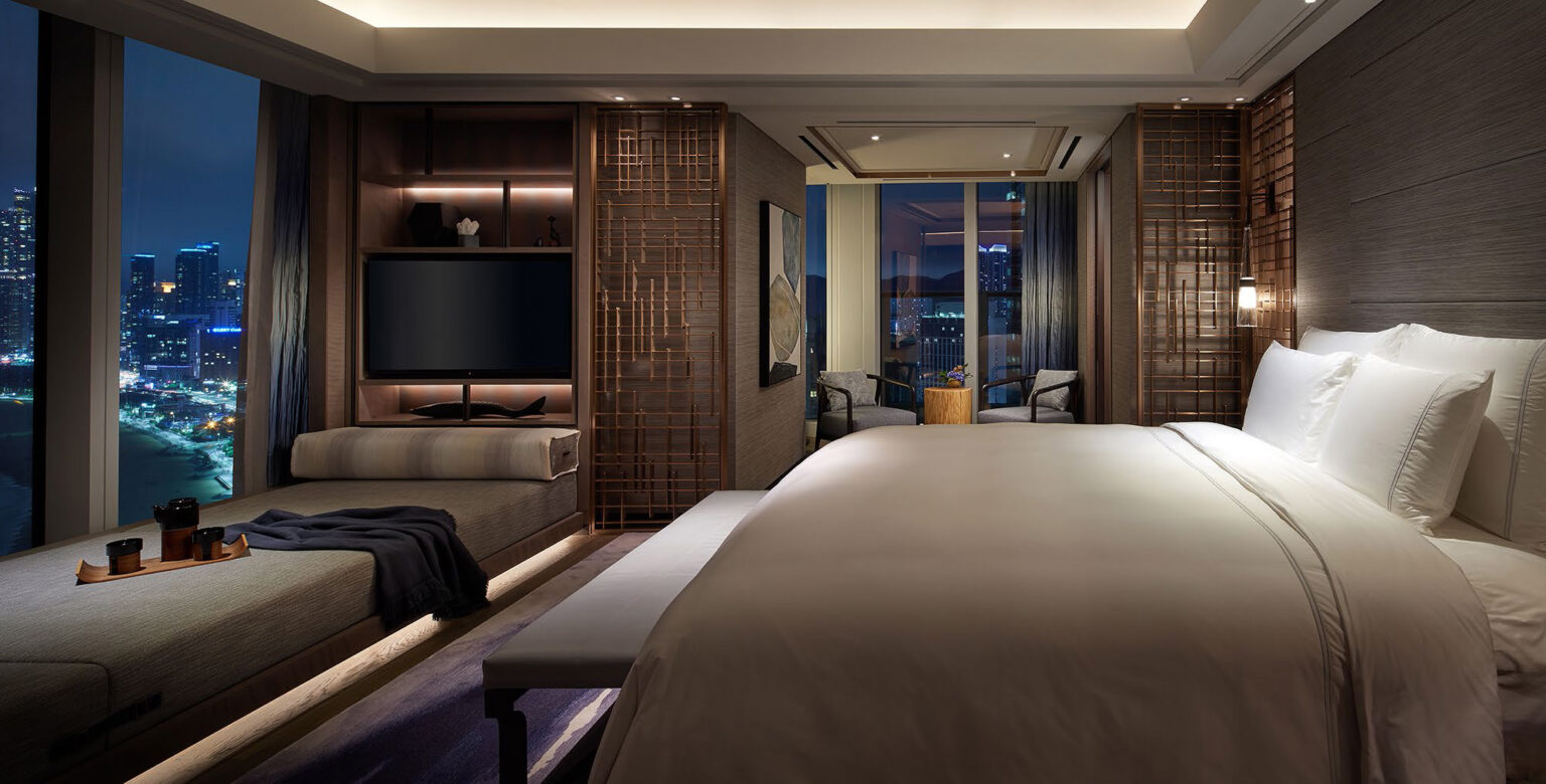 A luxurious suite with a large bed, sleek sofa, and a city view through floor-to-ceiling windows. The ambiance is elegant and tranquil.