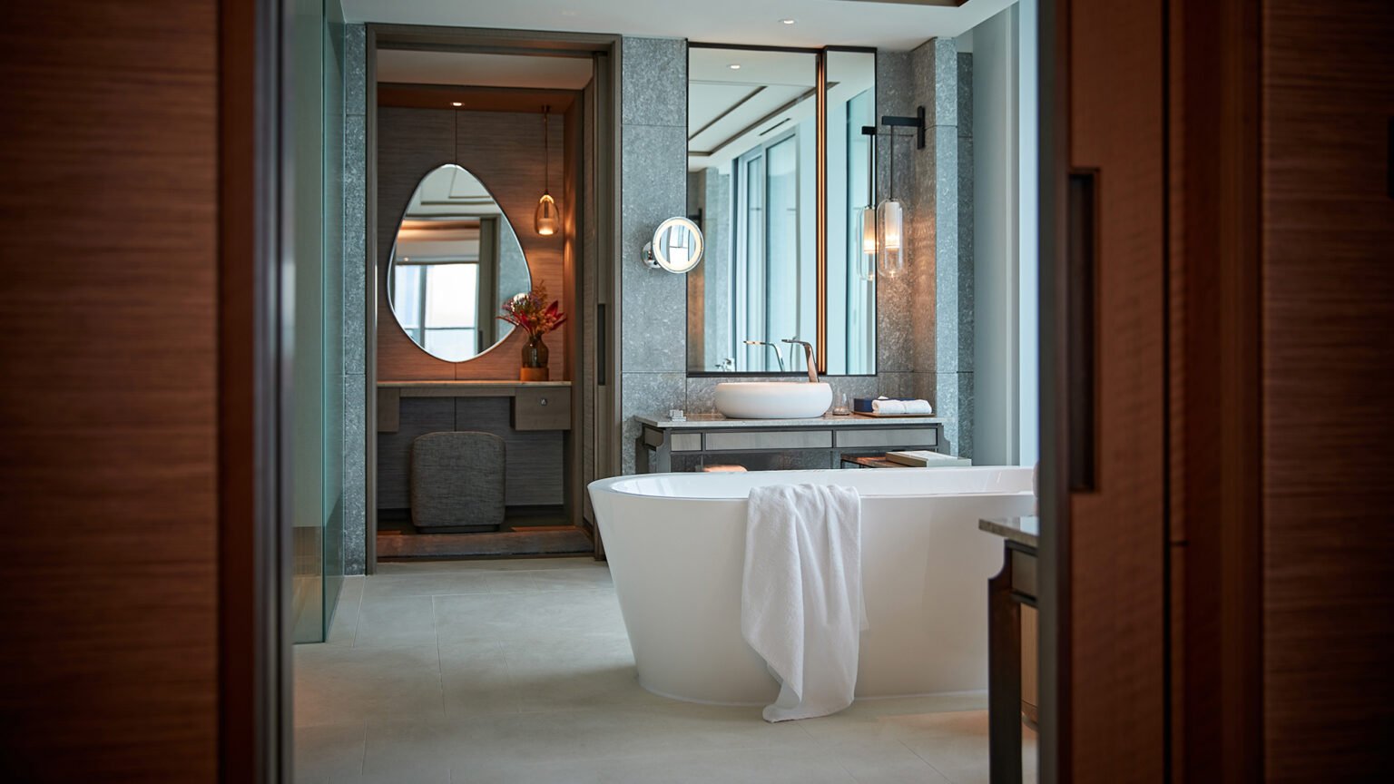 Luxurious bathroom featuring a freestanding white tub with a towel, modern fixtures, stone walls, an oval mirror, and a vase with red flowers, exuding serenity.