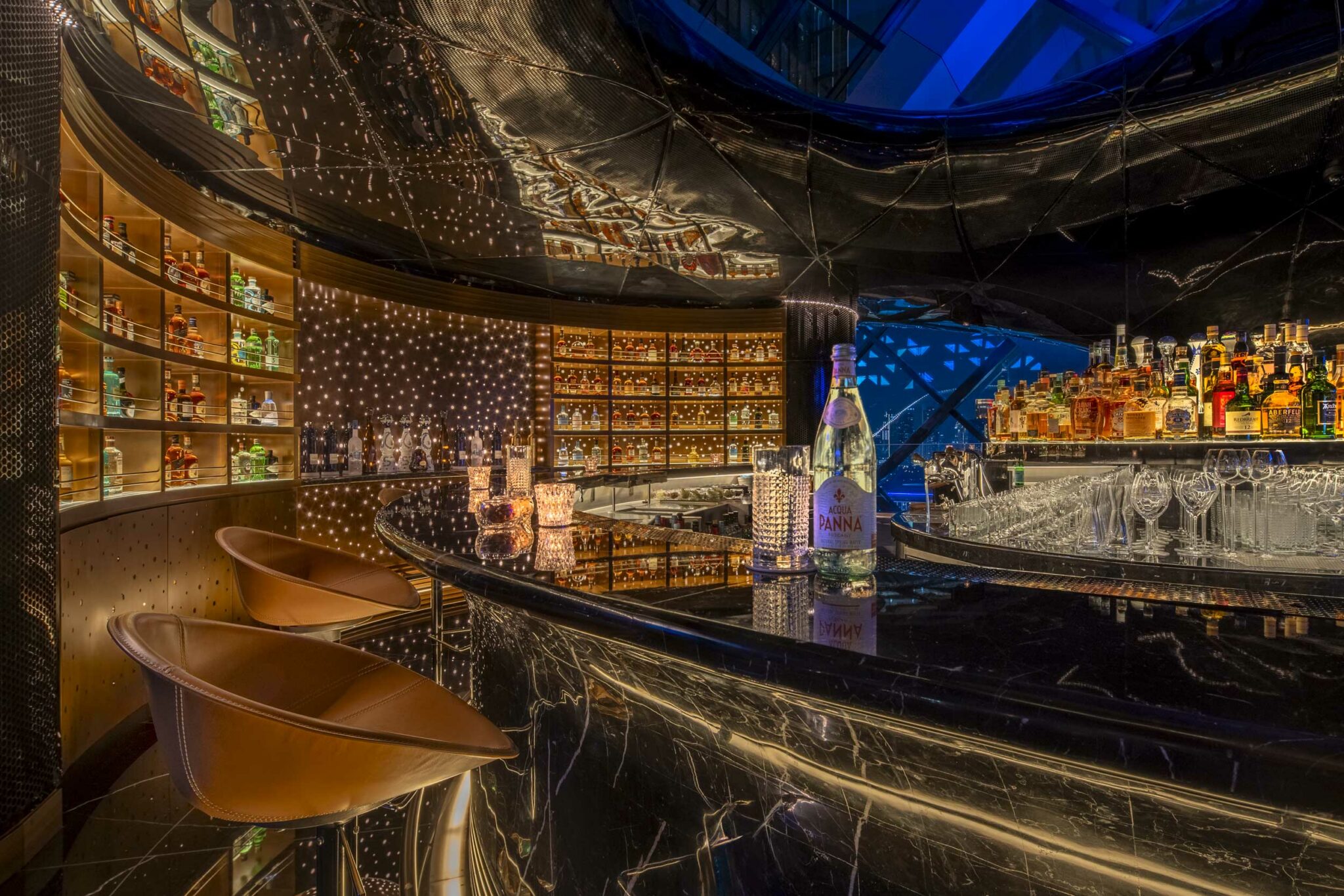 Luxurious bar interior with a sleek, black marble counter, leather stools, and shelves of illuminated bottles. Warm lighting creates a sophisticated ambiance.