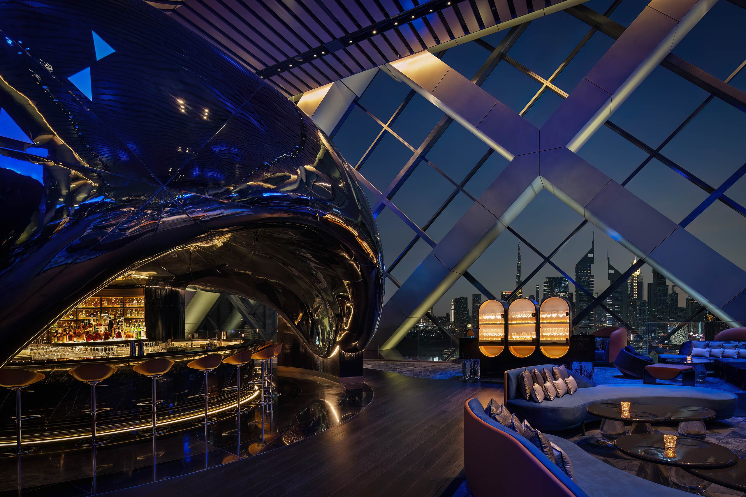 Futuristic bar with sleek black and metallic design, cozy seating, and glowing lights. Large windows showcase a Dubai's vibrant city skyline at night.