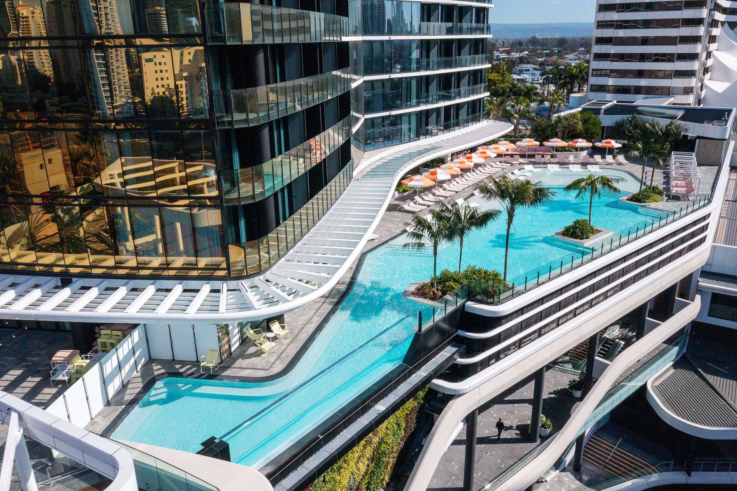 Aerial view of a modern hotel pool area with sleek architectural design, featuring a curving blue pool, palm trees, sun loungers, and orange umbrellas.