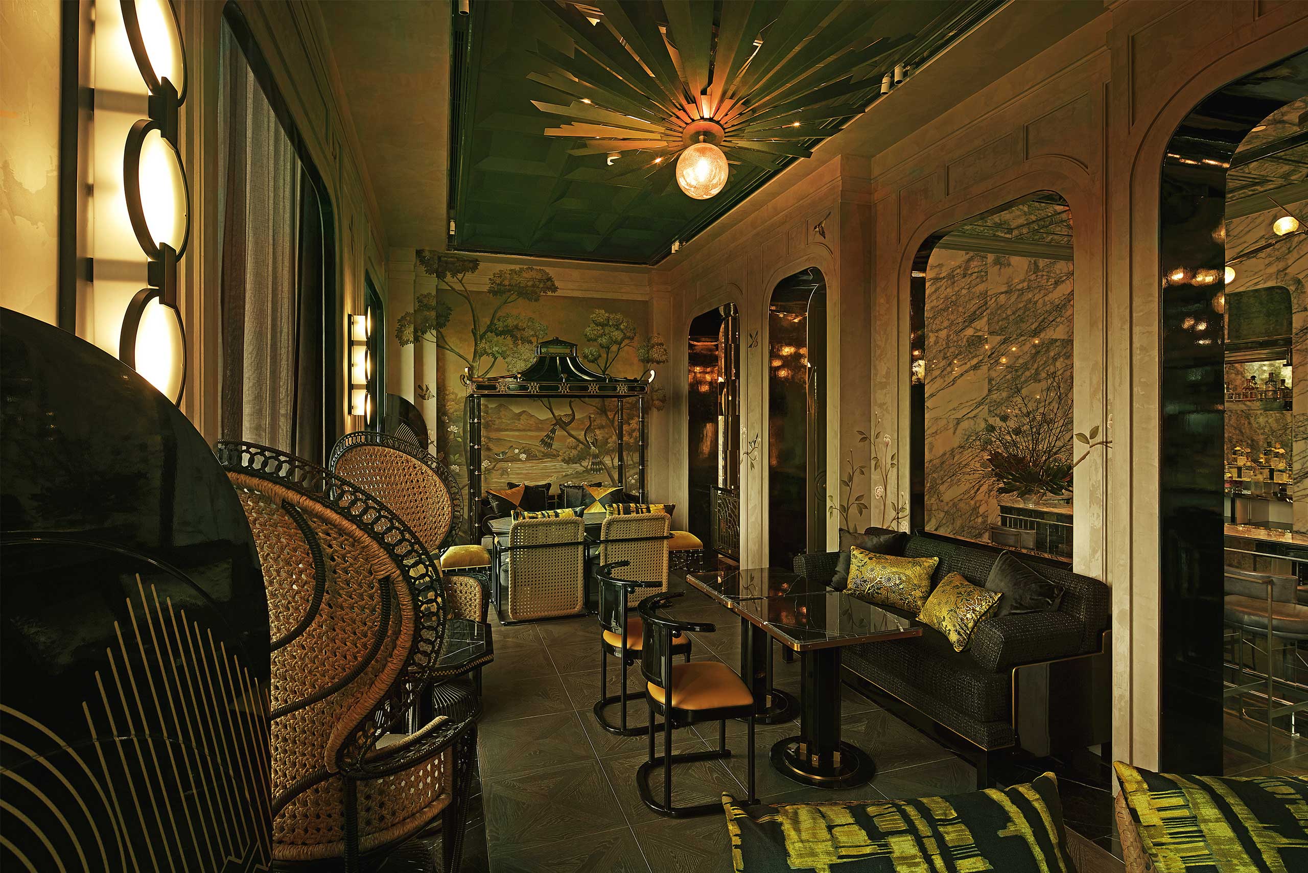 Elegant, dimly lit bar lounge with wicker chairs, plush sofas, dark tables, and a decorative green ceiling. A mural of trees and a large pendant light add charm.