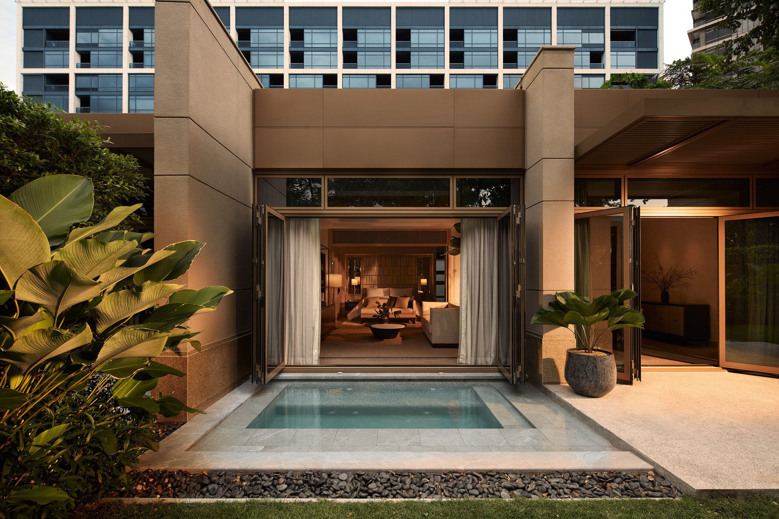 Outdoor view of a modern, luxurious apartment patio with a small pool, green plants, large open glass doors, and warm interior lighting visible inside.