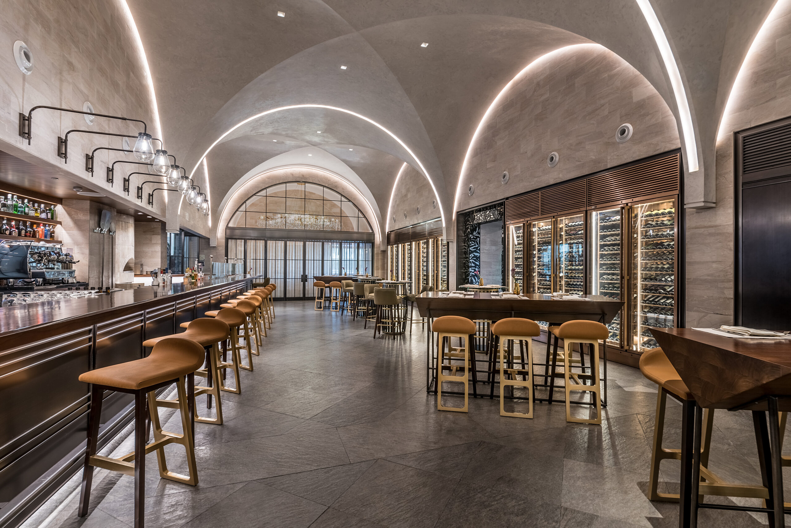 A modern bar interior with arched ceilings, warm lighting, and sleek design. Barstools line the counter, adjacent to tables and a wall of wine bottles. Cozy and inviting atmosphere.