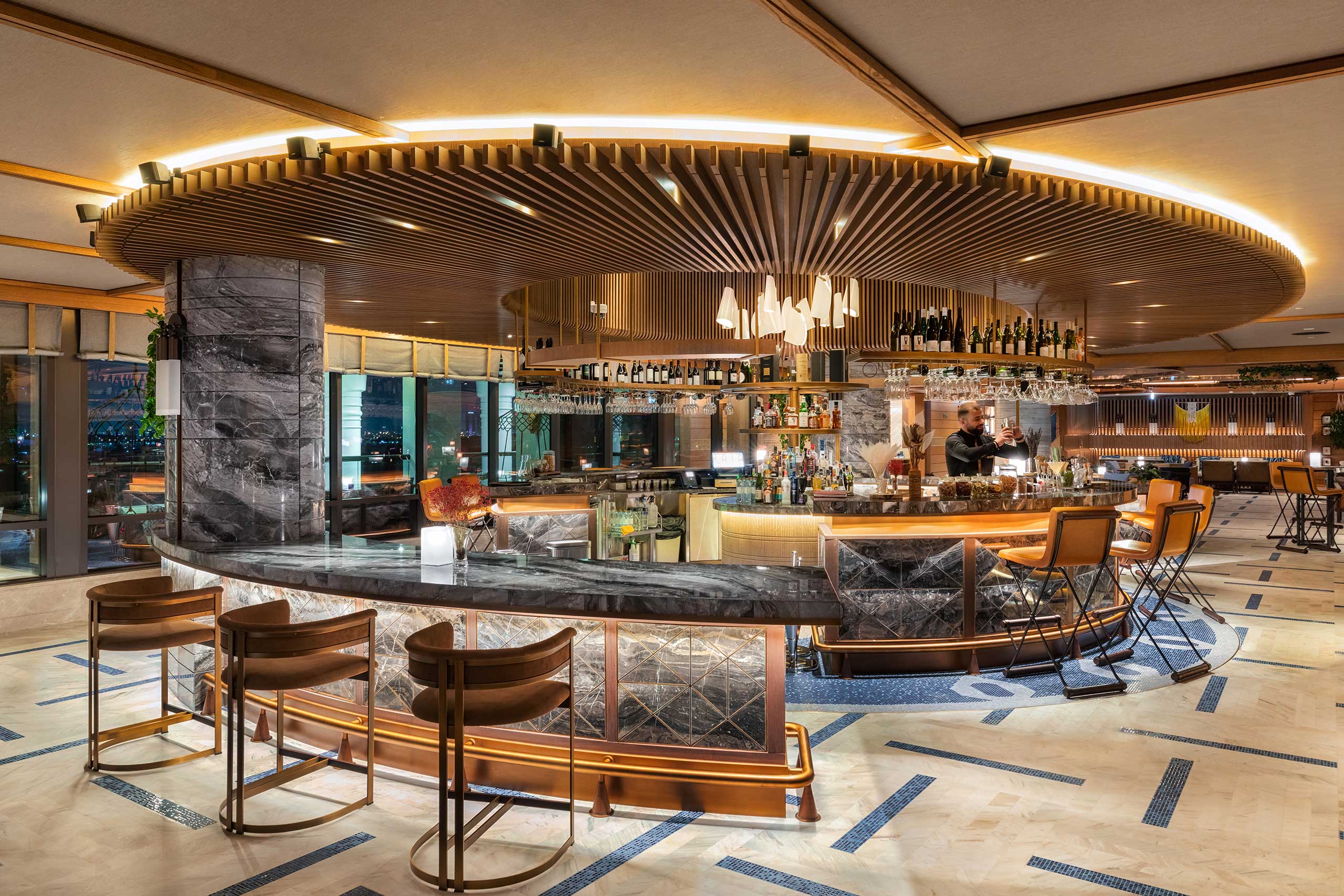 Modern, elegant bar with a circular marble counter, wooden accents, and a lit ceiling. Warm lighting creates a cozy ambiance, with stocked shelves and plush seating.