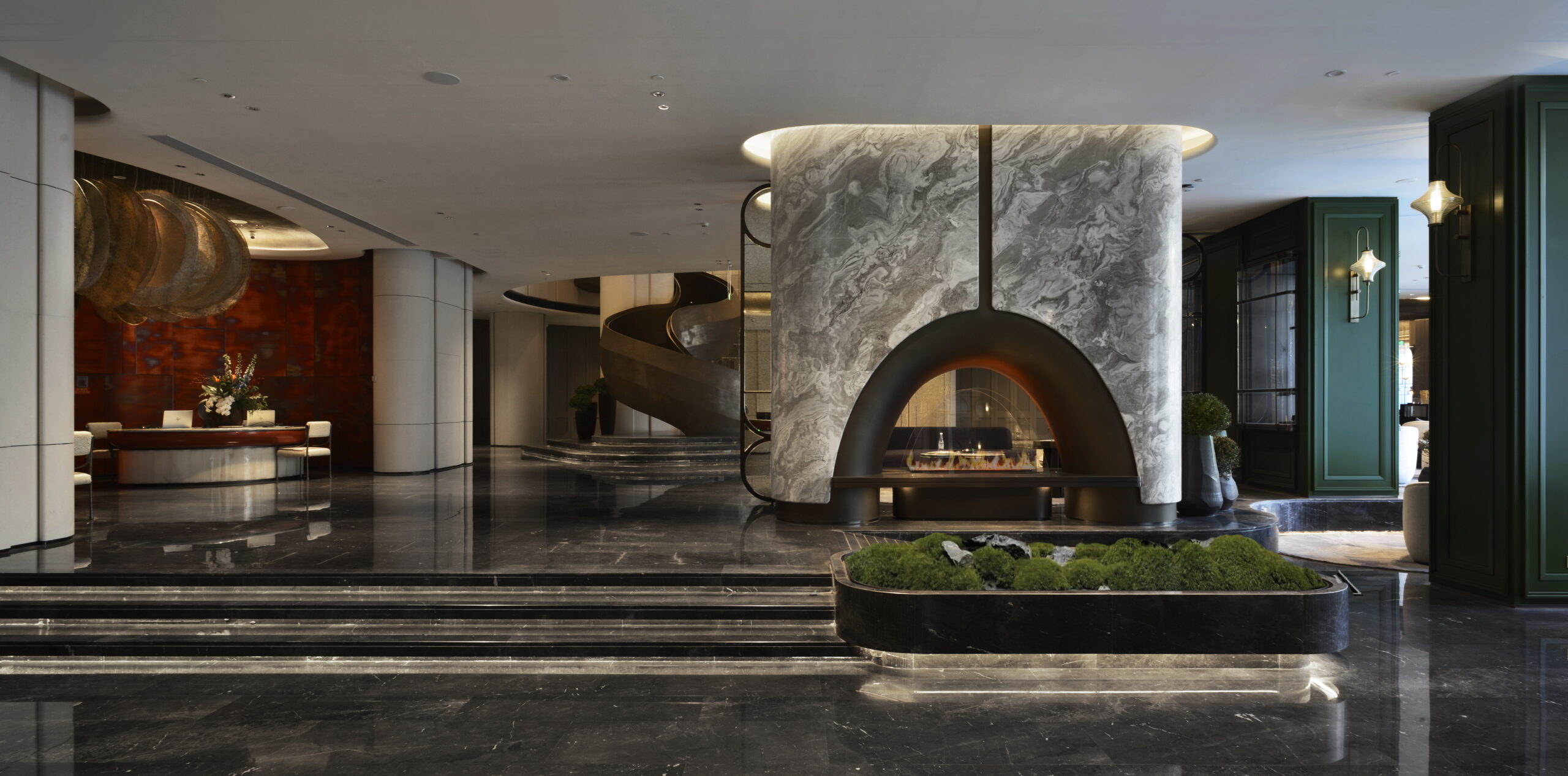 Luxurious hotel lobby with a central marble fireplace, sleek black floors, and a curved staircase. Decor is modern with plants and soft lighting, exuding elegance.