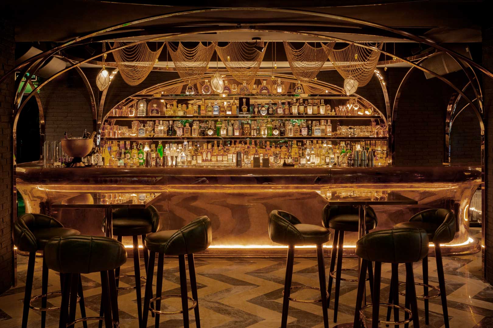 Luxurious, dimly lit bar with elegant shelving stocked with bottles. Ornate gold accents, plush green stools, and warm ambient lighting create an opulent atmosphere.