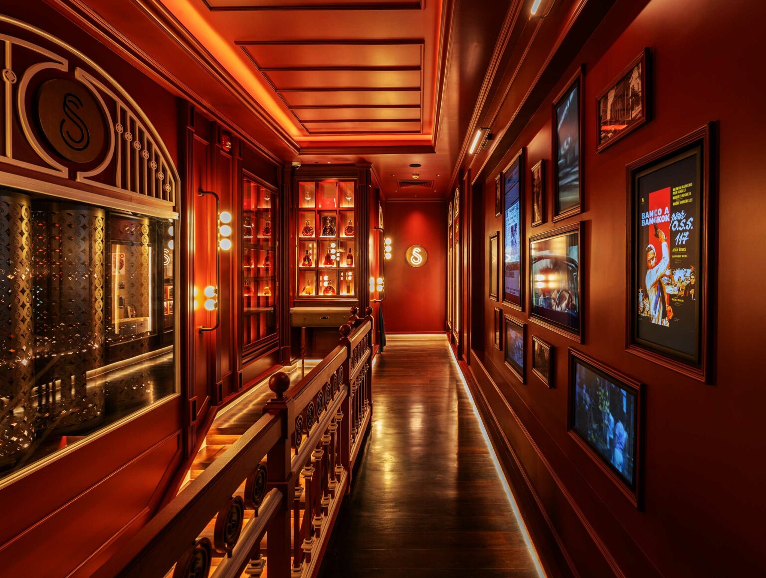 Warm, dimly lit hotel hallway with rich red walls and wooden accents. Shelves display bottles, while framed pictures and posters line the opposite wall, evoking elegance.