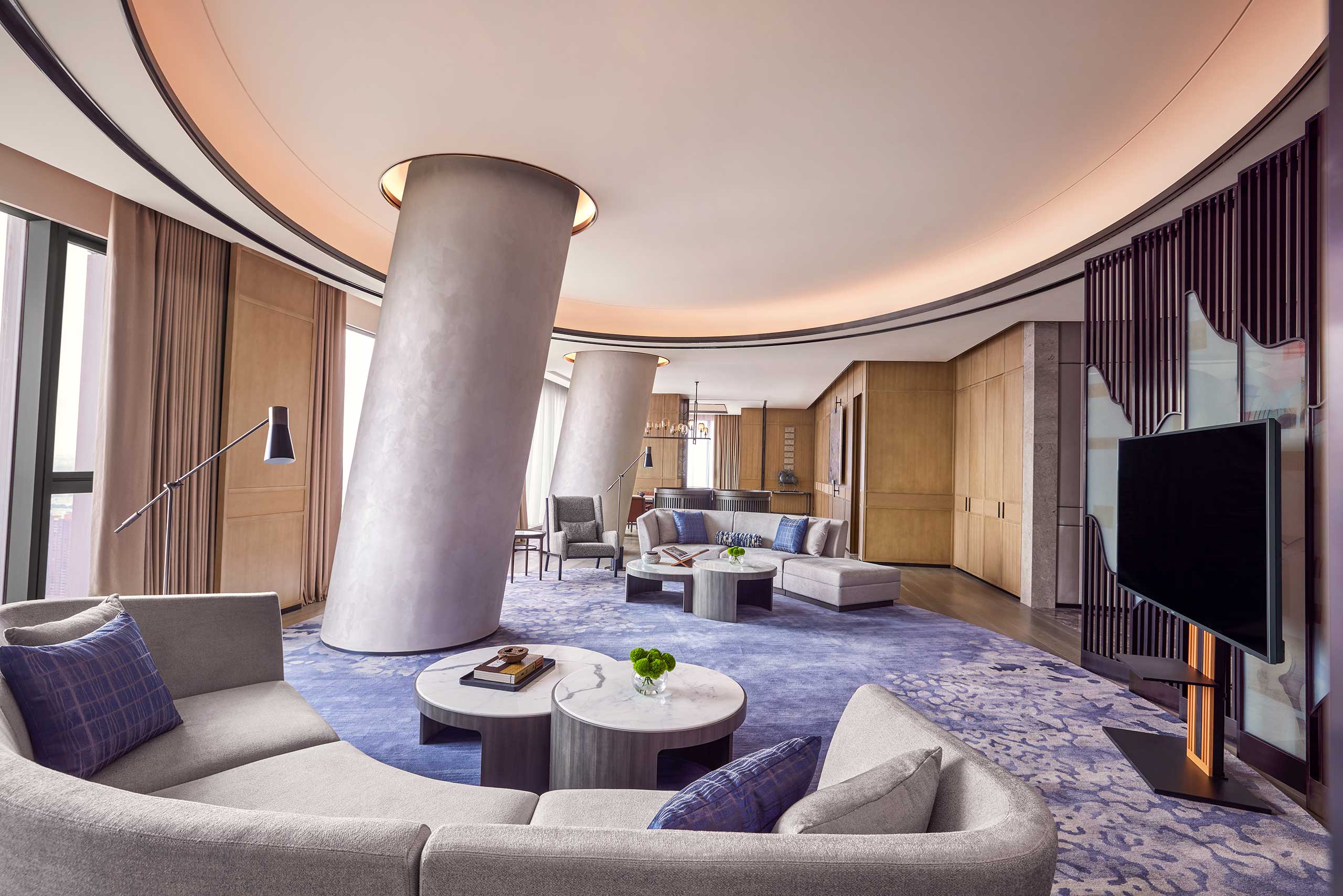 Spacious, modern living room with curving columns, plush grey sofas, and blue accents. Soft lighting creates a calm, sophisticated atmosphere.