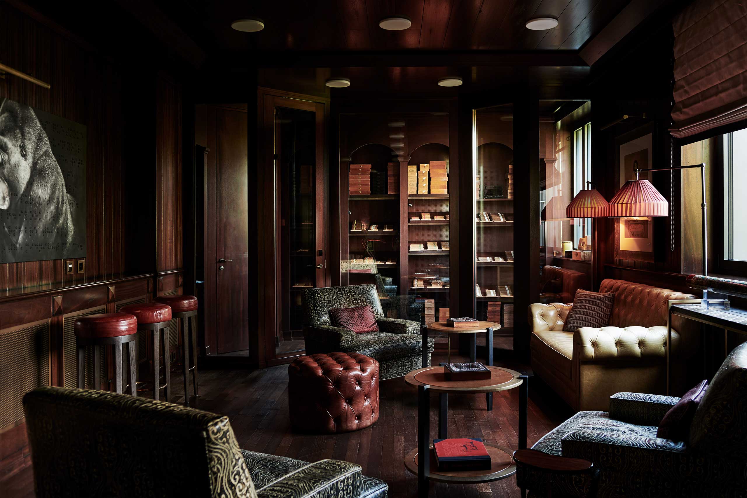 A cozy, dimly-lit hotel lounge with plush seating, a tufted leather ottoman, dark wooden walls, bookshelves, warm lighting, and a bear painting on the wall.
