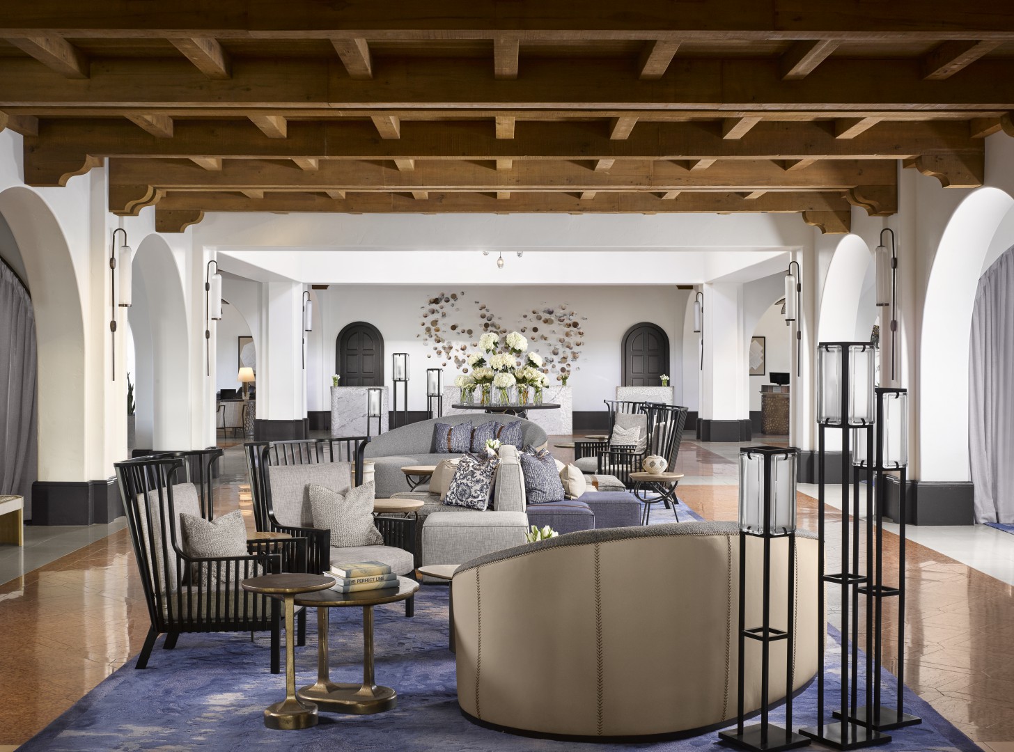 Elegant hotel lobby with plush sofas, decorative black chairs, and modern lamps on a blue rug. Arched doorways and floral centerpiece create a welcoming, luxurious ambiance.
