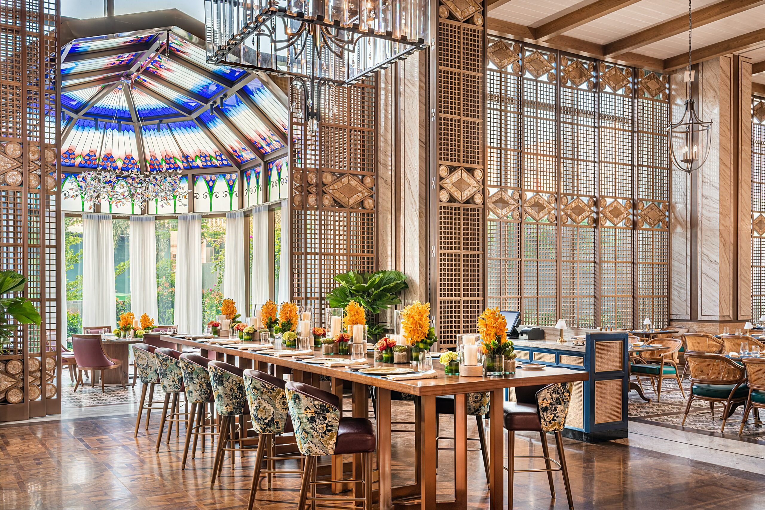 Luxurious resort restaurant interior featuring a long wooden table with floral chairs, vibrant yellow flowers, stained glass ceiling, and wooden lattice walls.