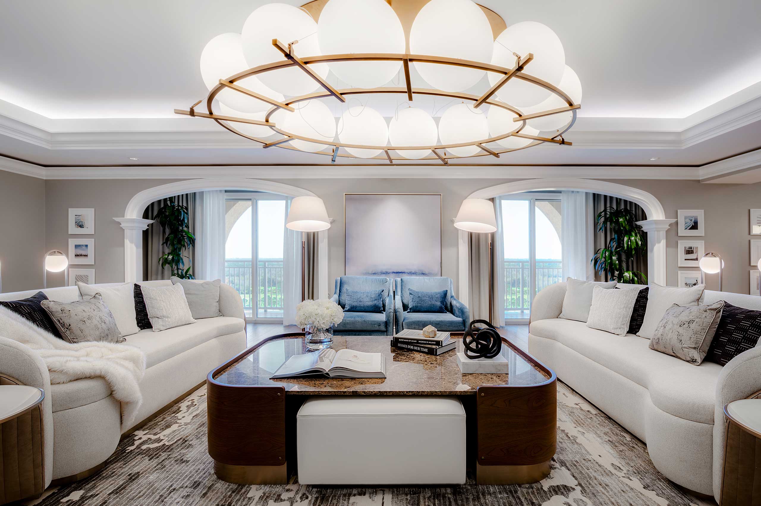 Elegant suite living room with two white sofas and blue armchairs facing each other. A large round chandelier hangs above a wooden coffee table with books and flowers, framed by large windows.