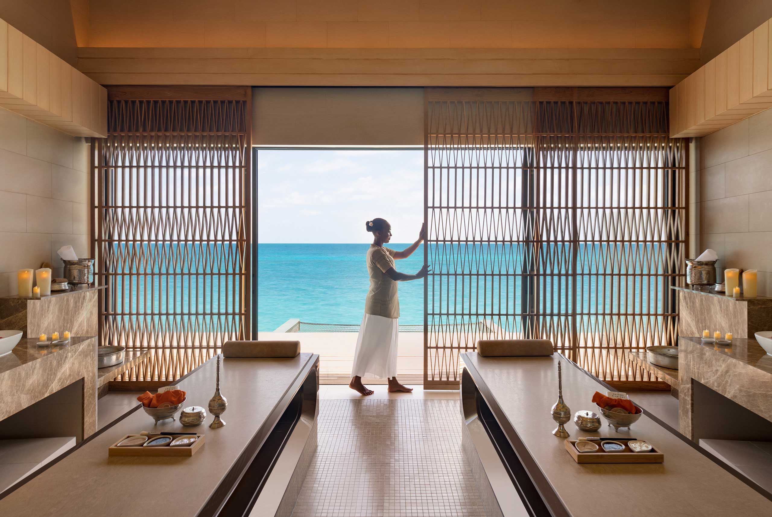 A serene spa treatment room opens to a turquoise ocean view. A person stands by sliding wooden doors, flanked by two massage beds and soft candlelight, creating a tranquil atmosphere.