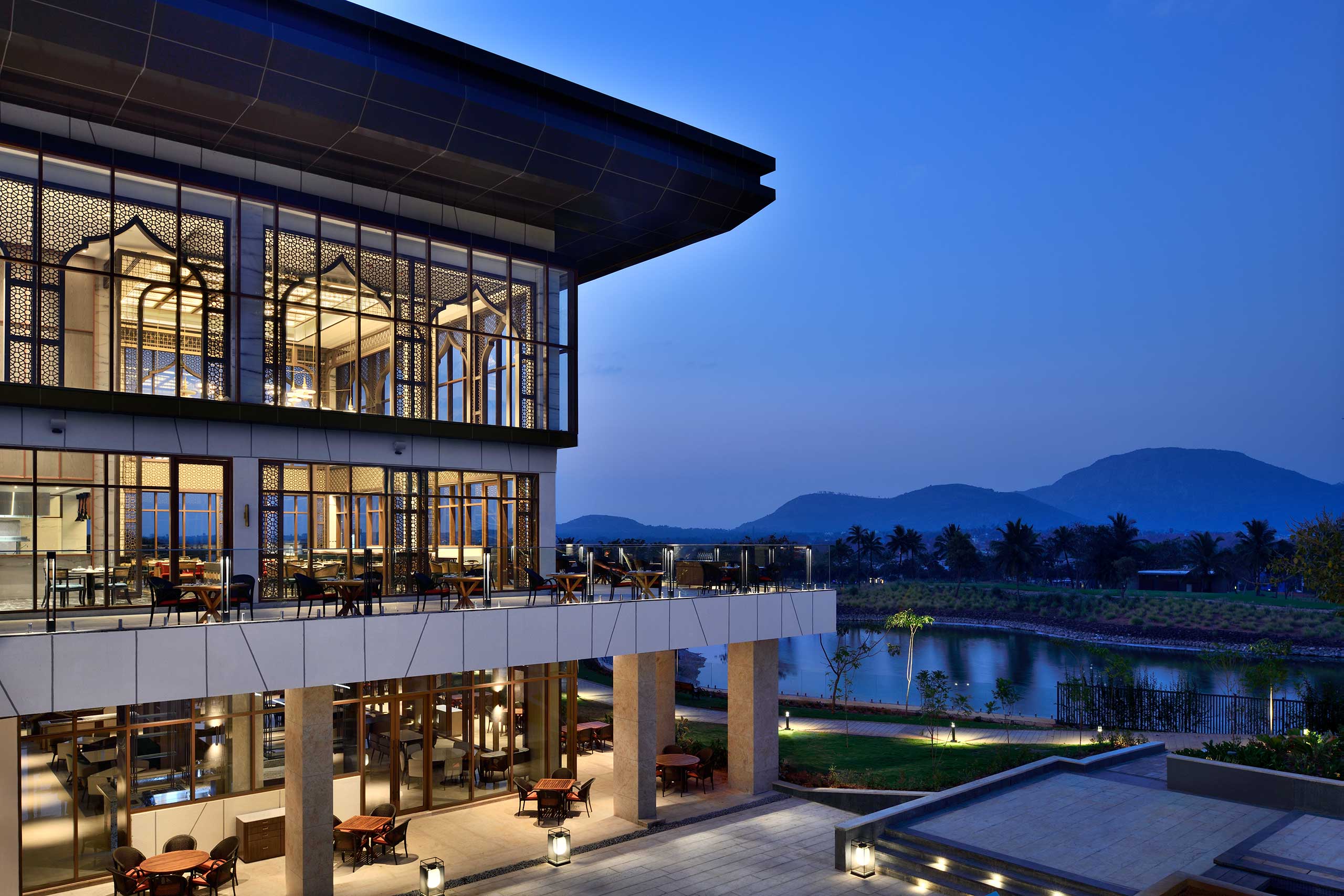 Modern building with large glass windows lit warmly at dusk. Overlooks a serene landscape with a pond and distant hills under a clear blue sky.