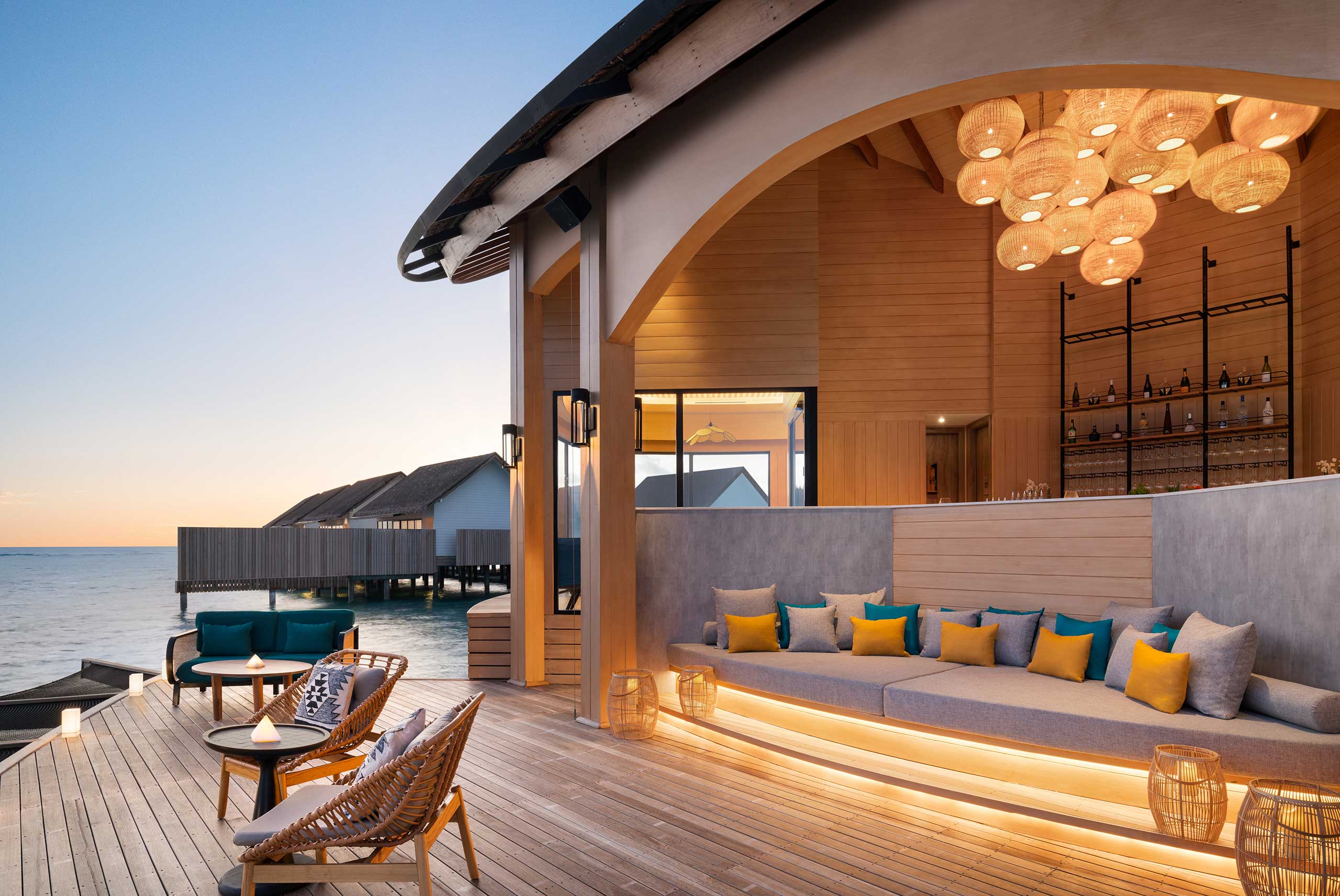 Luxurious beachfront bungalow lounge at sunset; overwater villas in background. Cozy seating with colorful cushions, warm lighting, elegant ambiance.